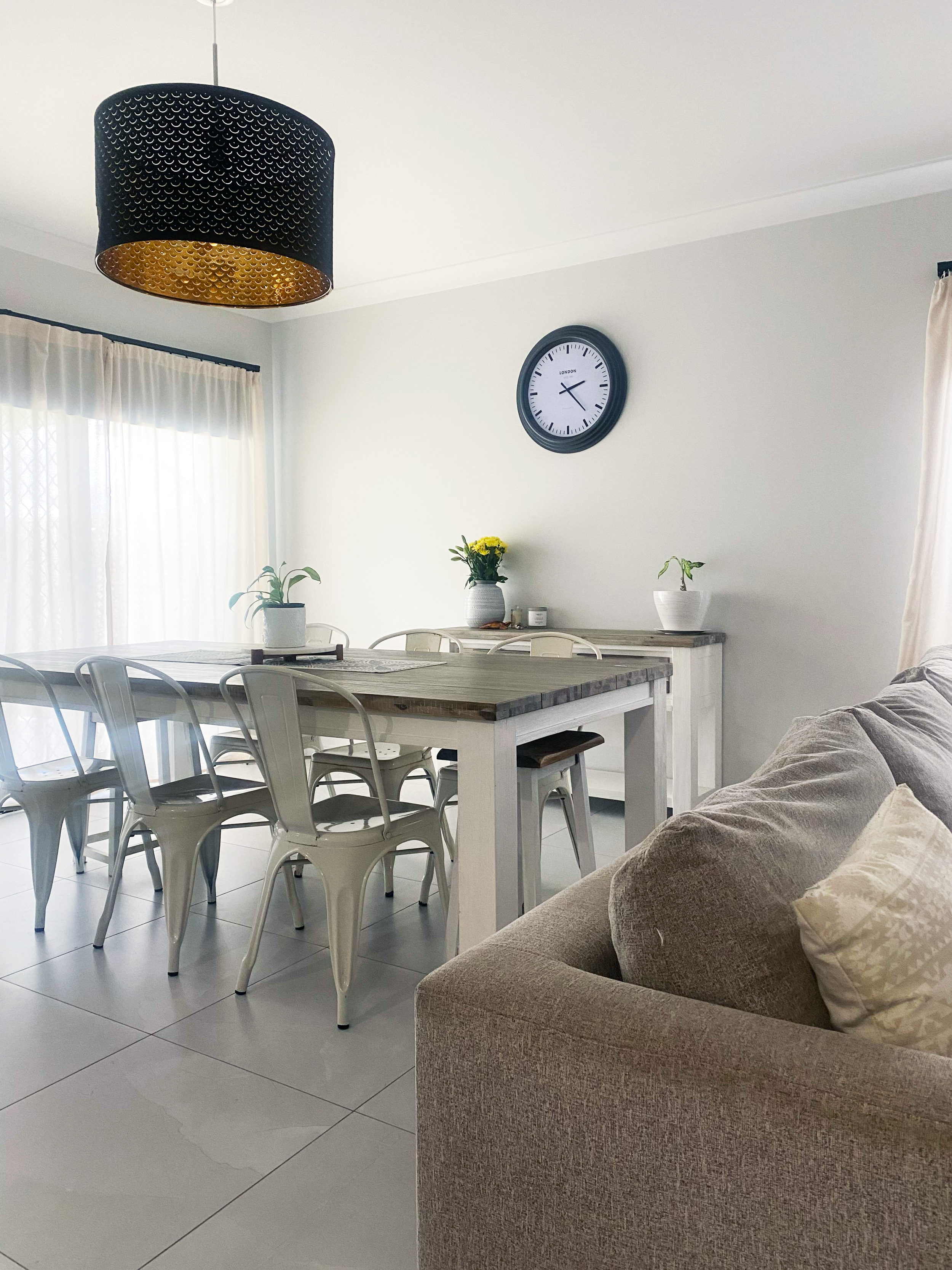 An image of the dining area in the Crestmead house. It has white walls, stylish white and wooden furniture, semi-transparent curtains and a large wall clock.