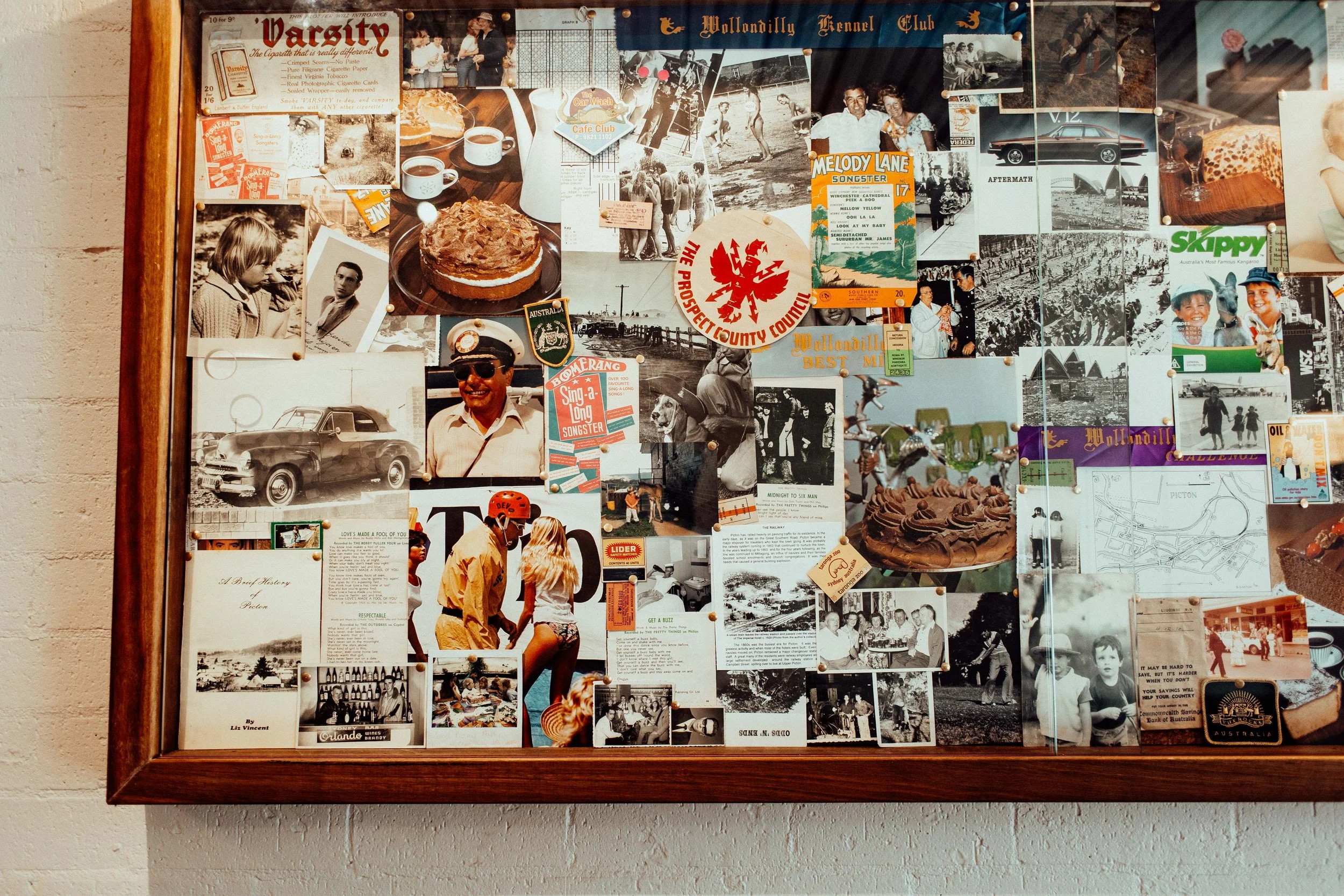 A bulletin board filled with various black-and-white and color photographs, posters, clippings, and memorabilia pinned to it. Some images include people, cars, and scenic views. There are also printed texts and tickets among the items. The frame of the board is wooden, and it is mounted on a white wall.