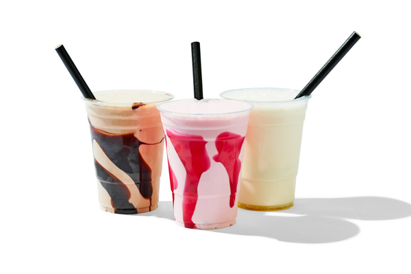 Three milkshakes with different flavors and toppings in clear cups with black straws, white background.
