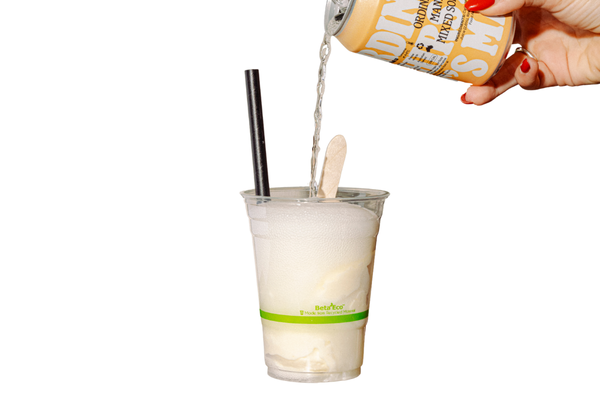 A hand pouring a can of orange soda into a cup of vanilla ice cream with a straw and a wooden stir stick.