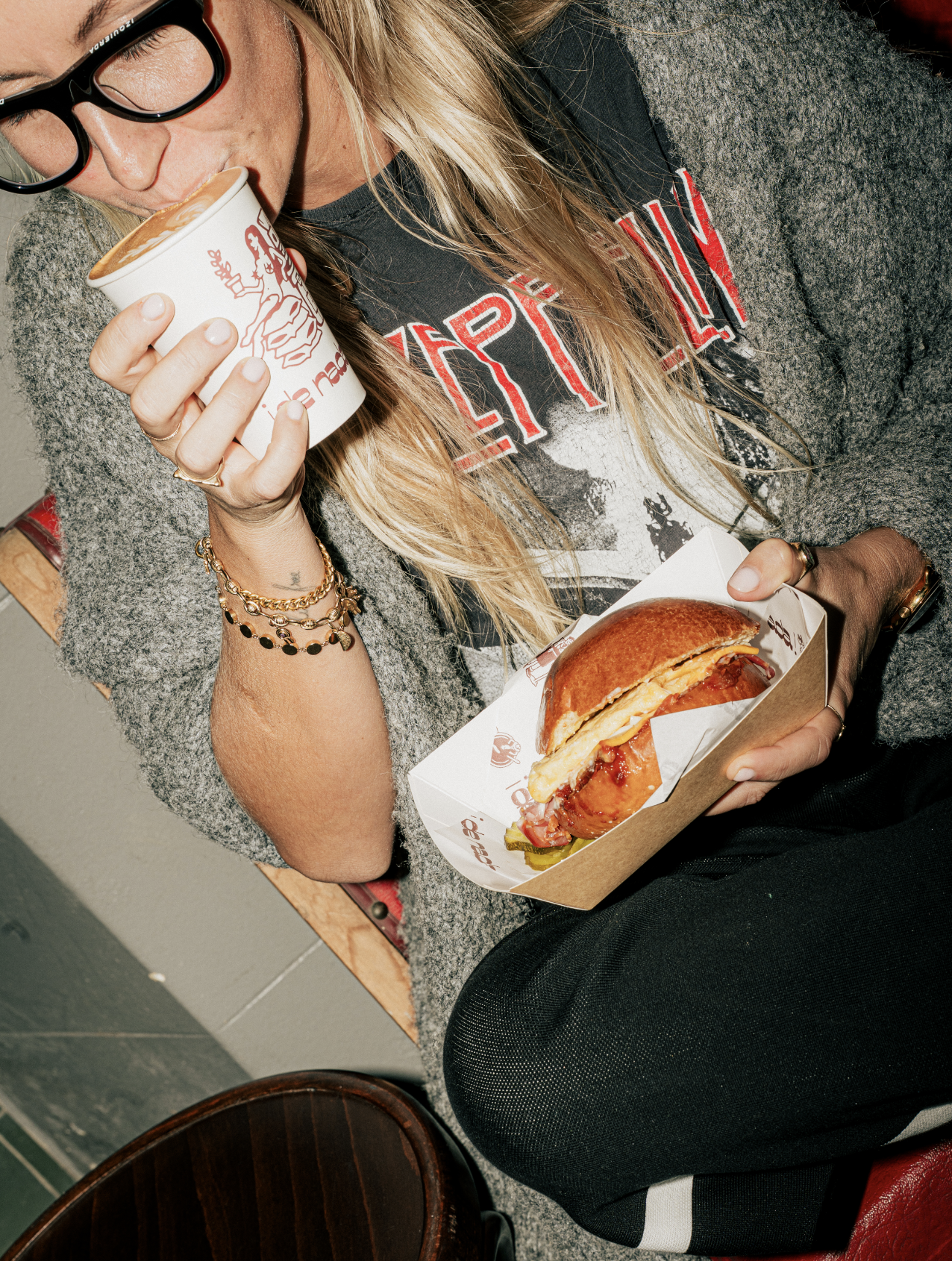 A woman with blonde hair, wearing glasses, a graphic t-shirt, and a gray sweater, sitting on a chair, holding a cup of coffee with a logo, and eating a sandwich in a small paper tray.