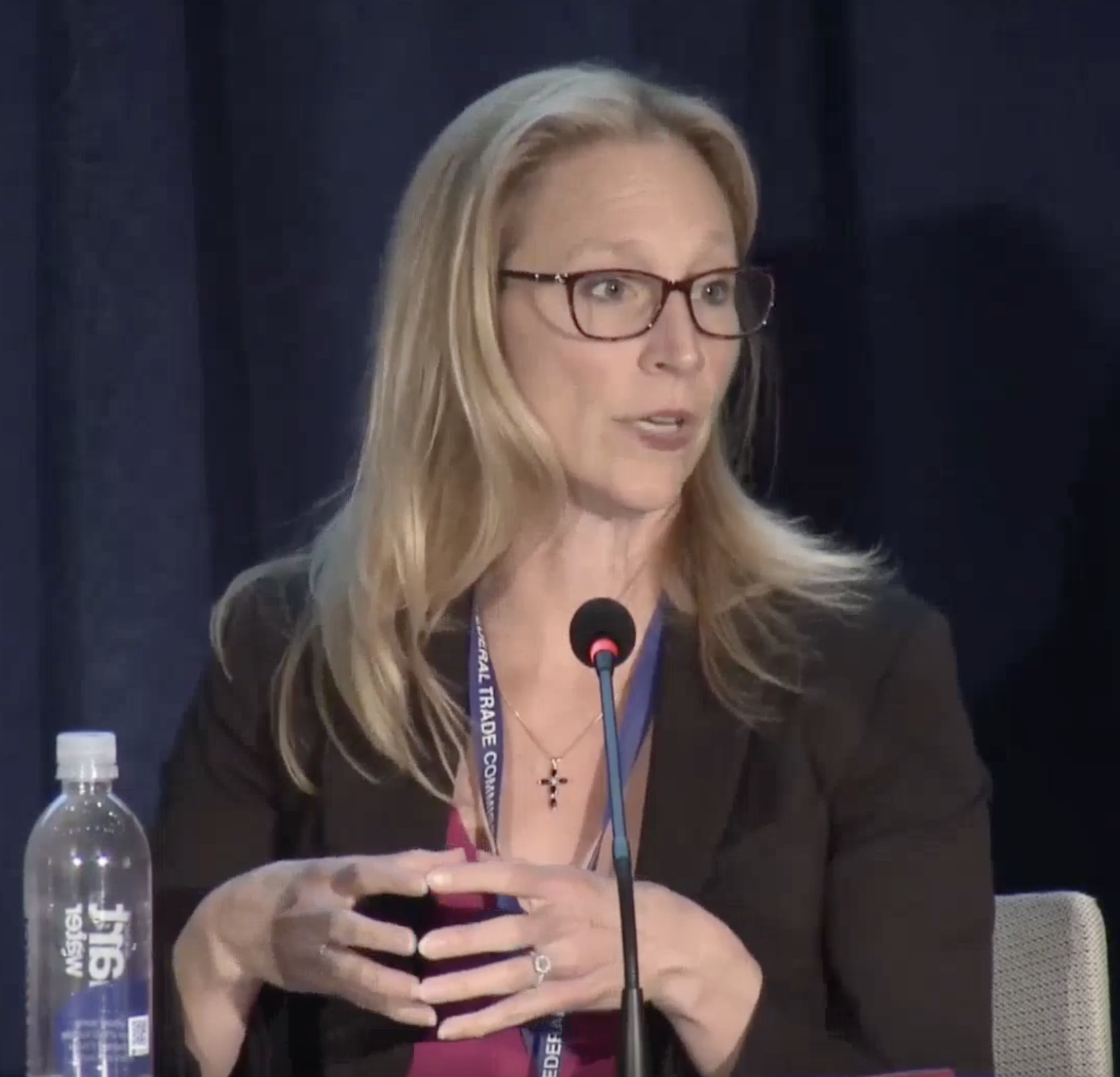 A woman with blonde hair and glasses speaking at a conference, sitting at a table with a microphone and a water bottle.