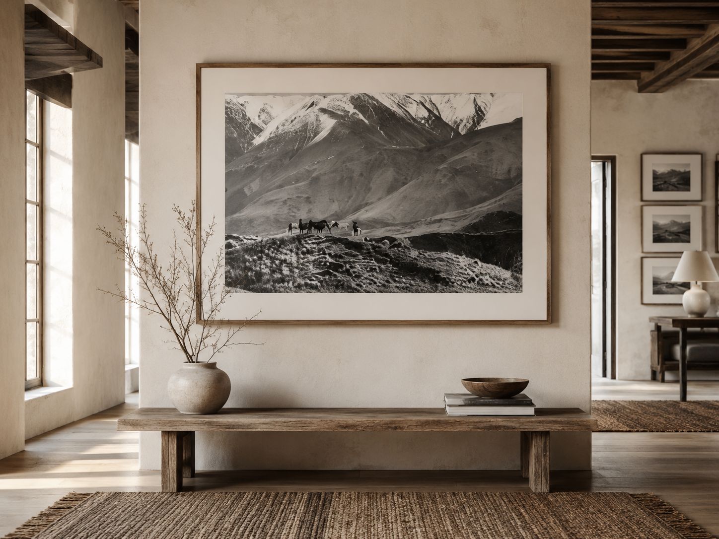 Decorative living room with large mountain landscape black and white framed photo, vase with branches, wooden bowl, stacks of books, linen curtains, wooden ceiling beams, and natural light.