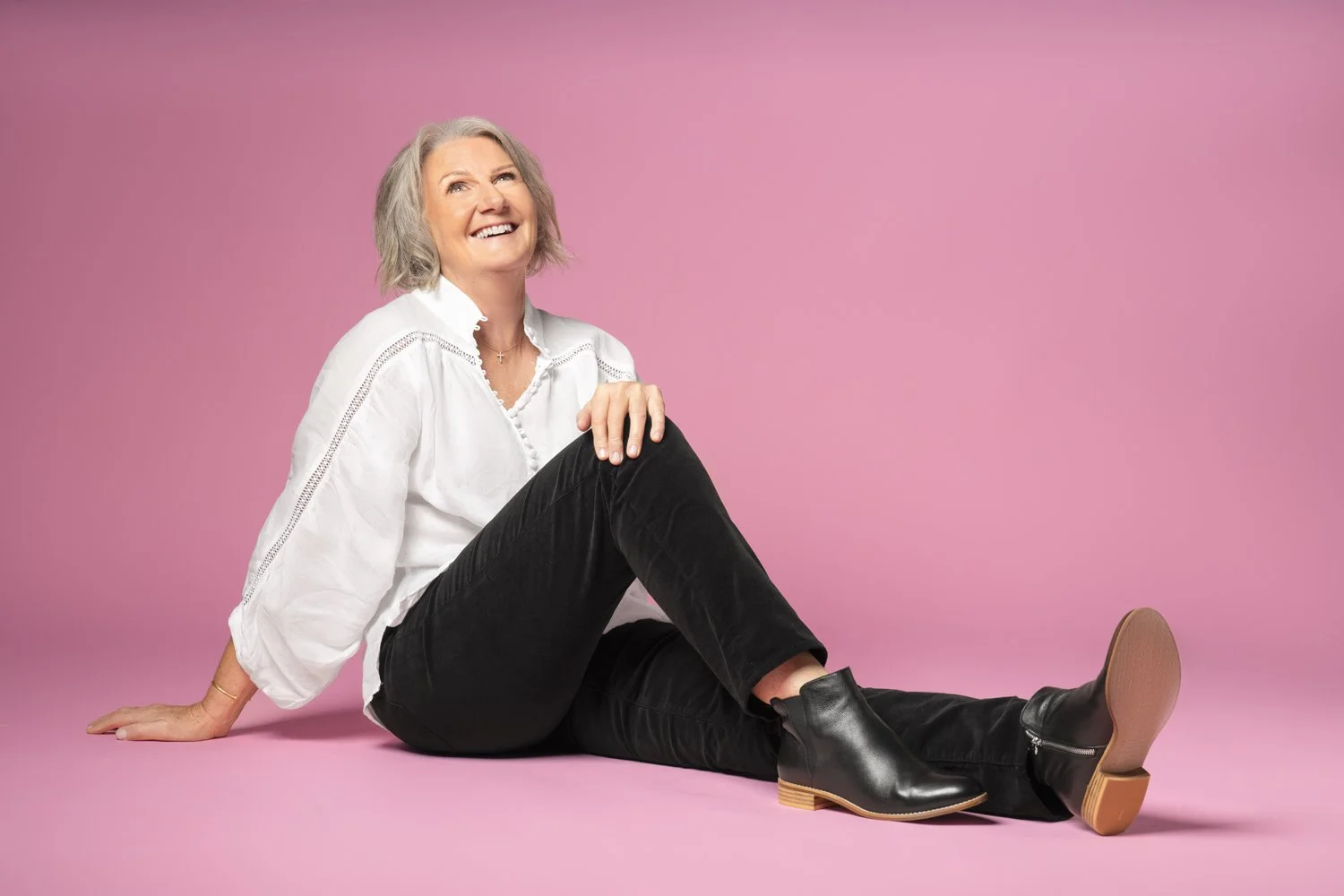 Diane Armstrong laughing, seated on the floor against a pink backdrop