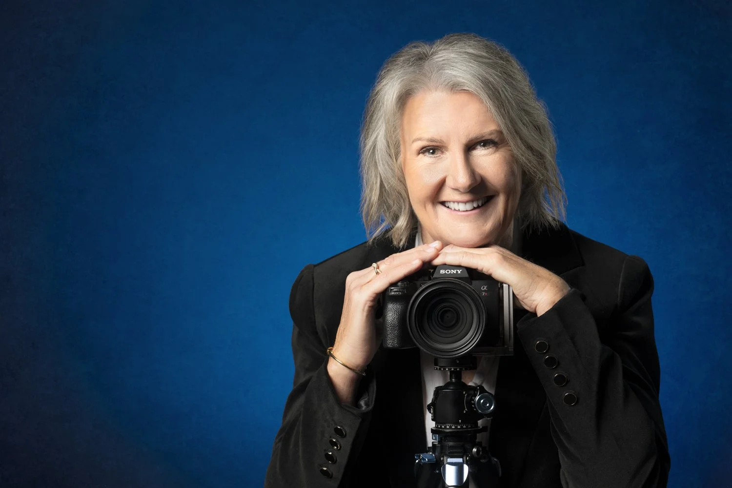 Diane Armstrong smiling behind her camera on a tripod, against a deep blue backdrop