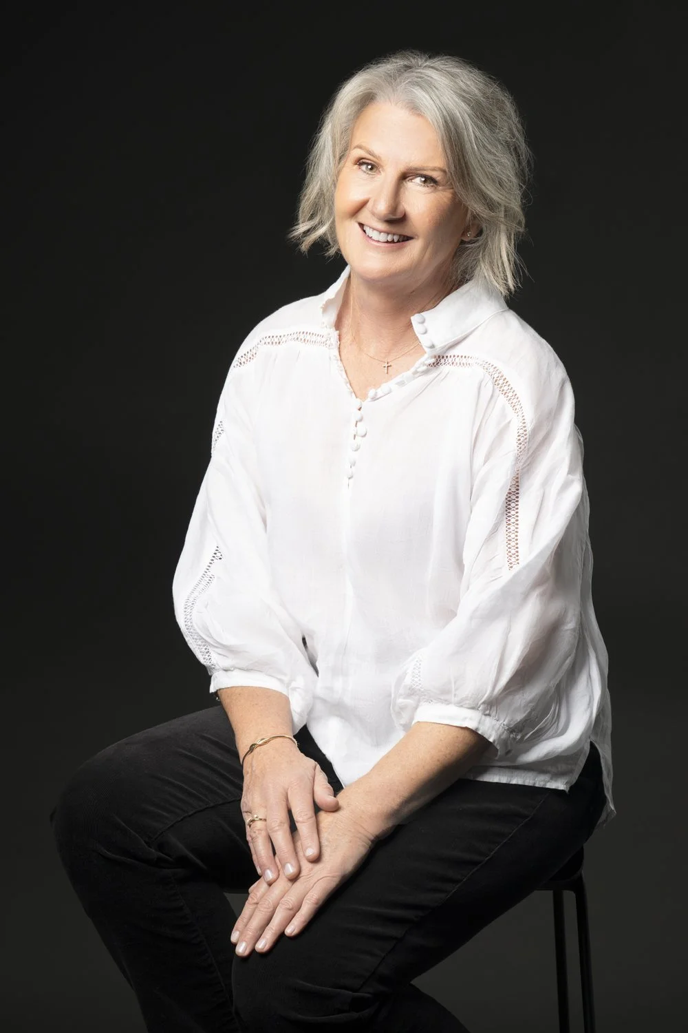 Diane Armstrong seated in a white shirt against a dark backdrop, smiling