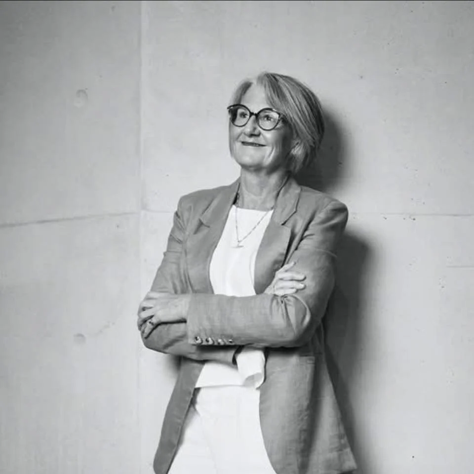 Black and white photo of a woman with glasses, short hair, wearing a blazer and t-shirt, standing with arms crossed and leaning against a wall.