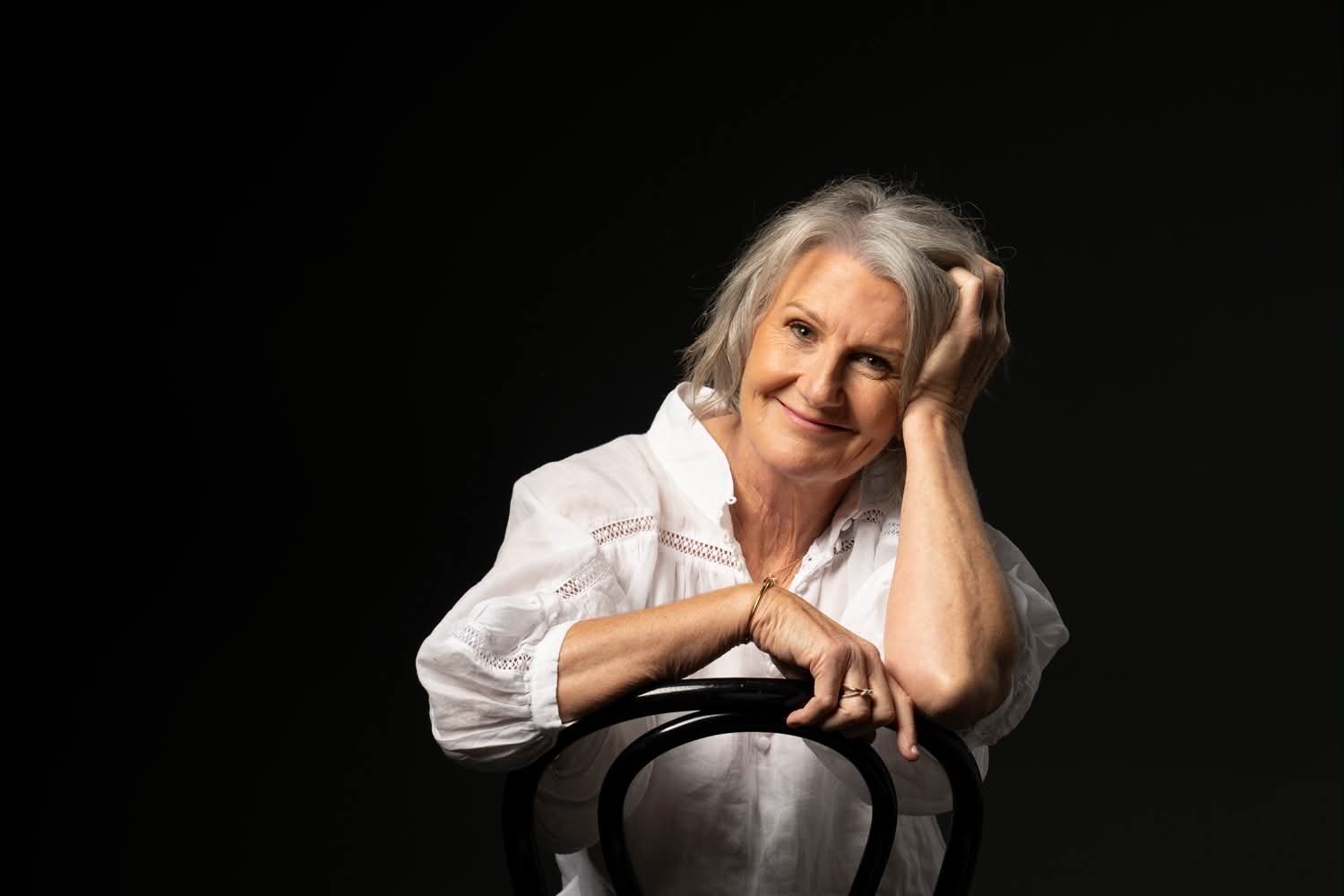 A senior woman with gray hair, smiling, resting her head on her hand, wearing a white blouse, posing against a black background