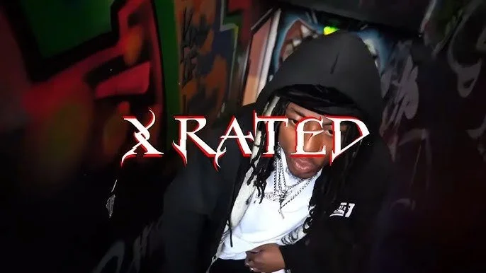 A person with long dreadlocks and glasses wearing a hoodie and white shirt, with the words 'X RATED' in large, stylized red and white text overlayed on the image, in a room with colorful wall art.