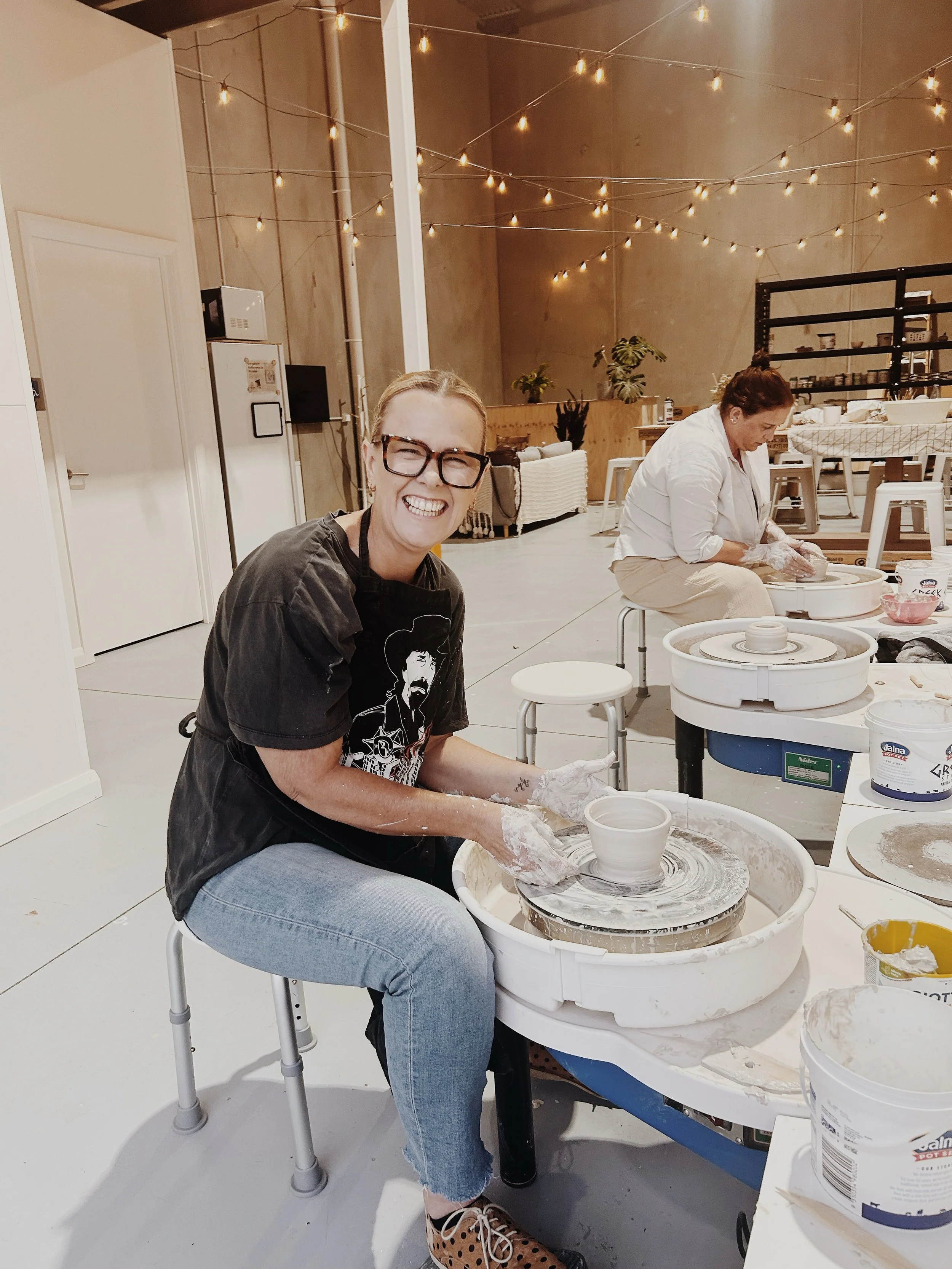Woman smiling at the camera while making pottery on a wheel in a studio, with another person working in the background, and string lights hanging from the ceiling.