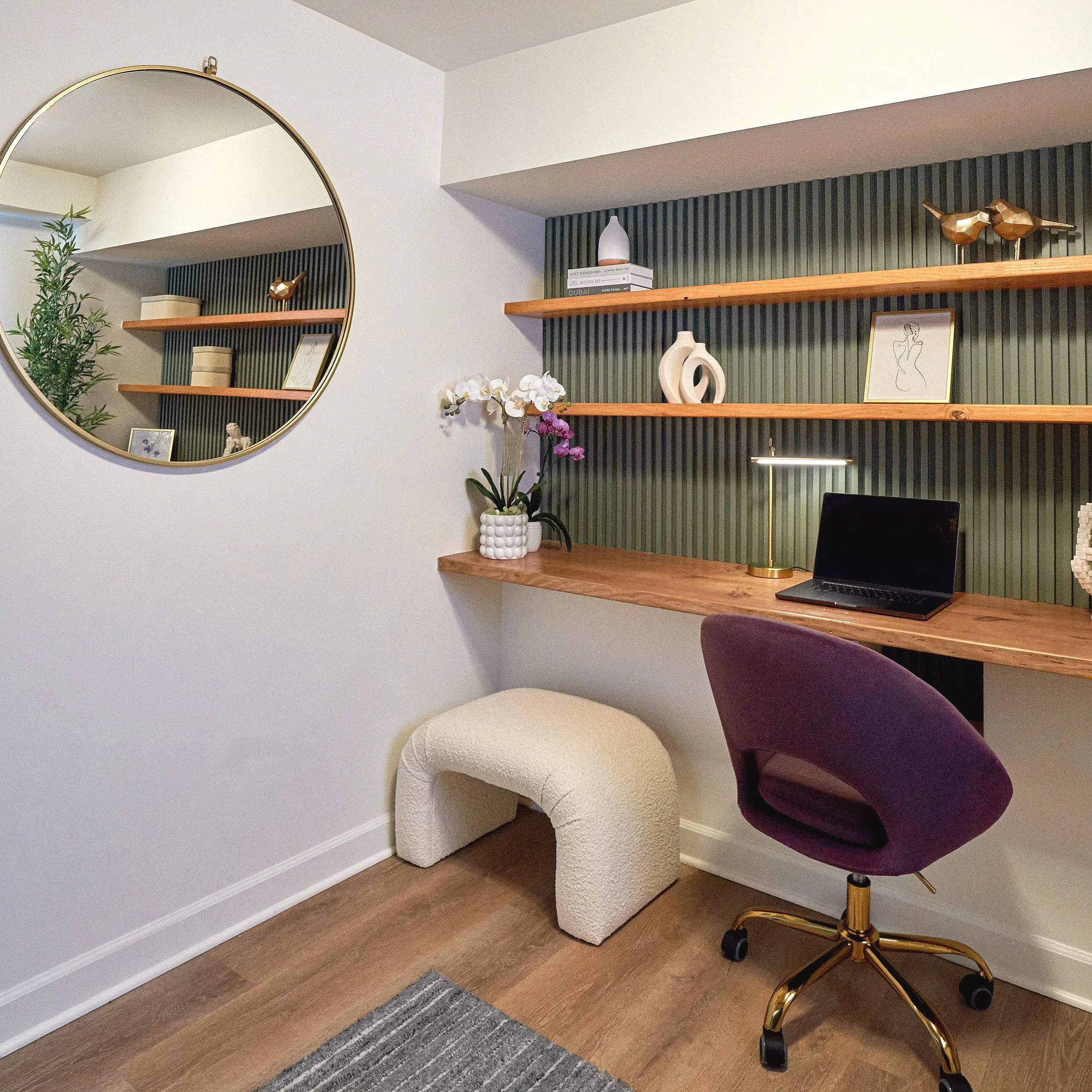A modern home office nook is built into a recessed wall, featuring a floating wooden desk and two matching wood shelves mounted against a dark green vertical slat accent panel. The shelves are styled with decorative objects including books, sculptura