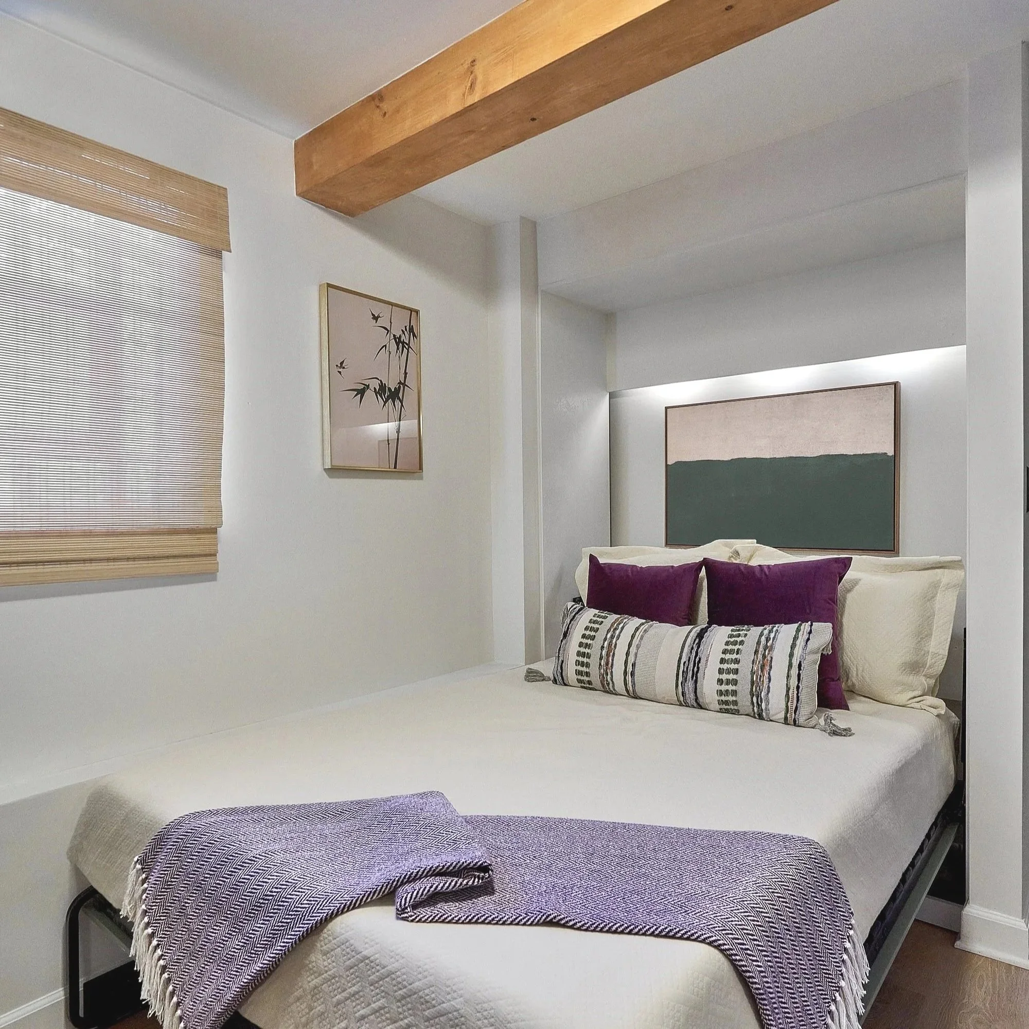 A modern bedroom features a wall-mounted Murphy bed lowered into a recessed alcove, styled with neutral bedding and layered accent pillows in cream, patterned fabric, and deep purple. A folded lavender throw blanket rests across the foot of the bed. 