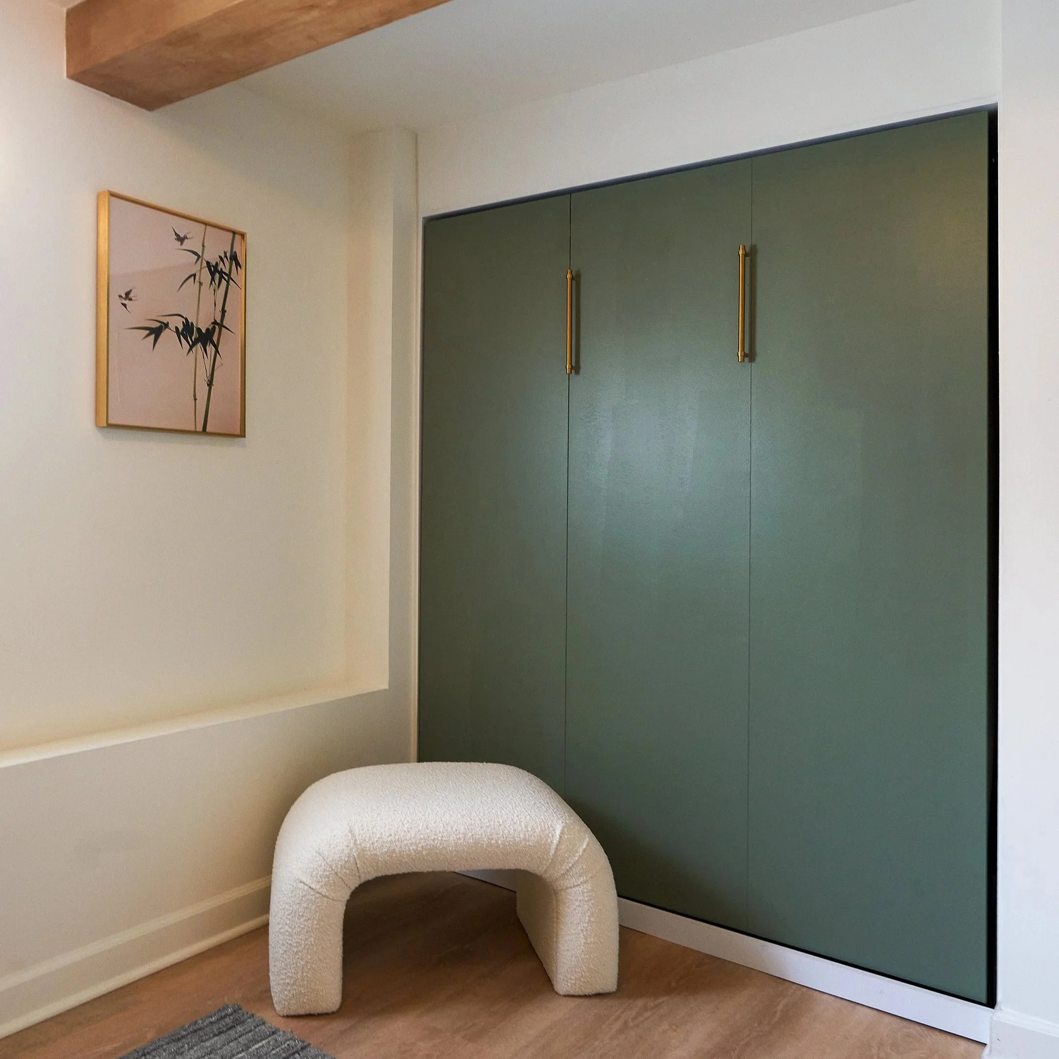 A modern multipurpose room features a closed Murphy bed concealed behind sleek green cabinet panels with long vertical brass handles. The cabinetry sits within a recessed wall beneath a natural wood ceiling beam that adds warmth and architectural cha