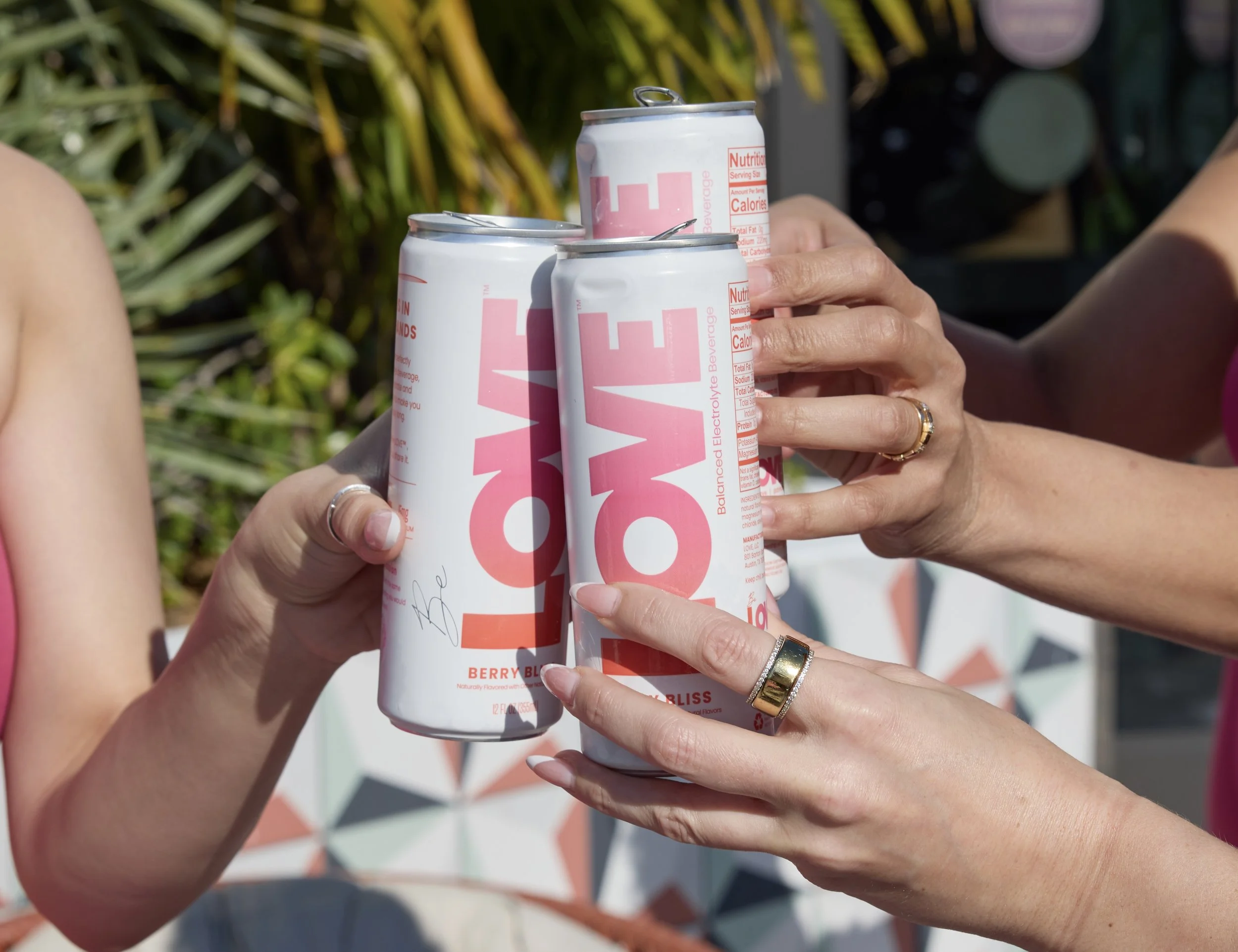 Three people holding and clinking cans of Love beverage with pink and white labeling, outdoors with bright sunlight.