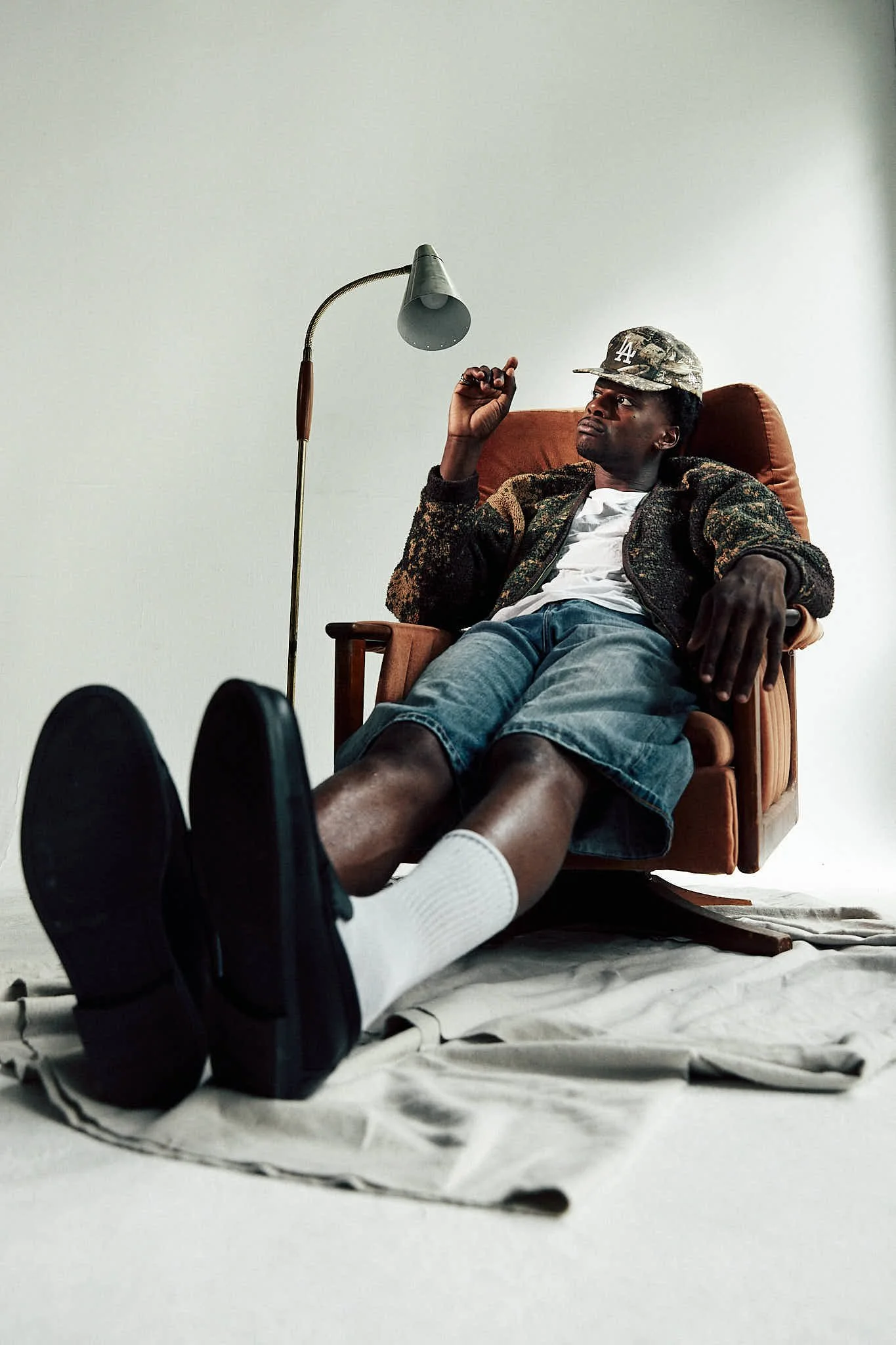 A man seated in an armchair with his feet stretched out on the floor, wearing a Los Angeles Dodgers cap, a patterned jacket, white t-shirt, and denim shorts, looking upward, with a vintage floor lamp nearby, against a plain wall background.