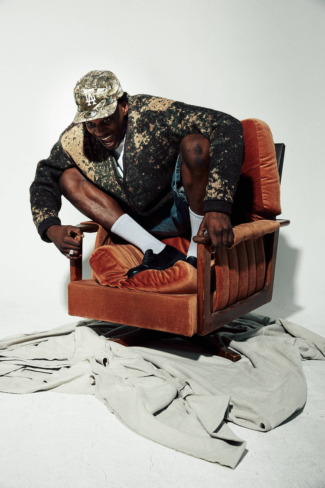 A man with a baseball cap, patterned jacket, and shorts crouches on a vintage orange chair with a wooden frame, smiling and leaning forward.