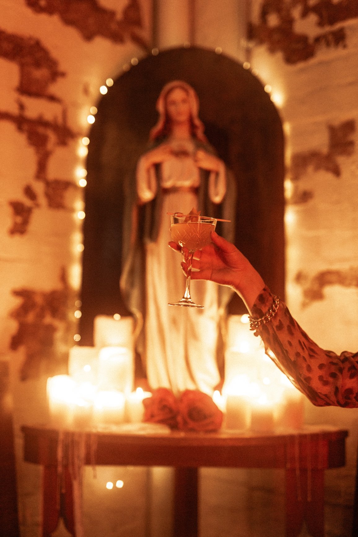 An image edited to be a yellow and red hue, a hand is just in frame, holding a cocktail in front of a religious altar.