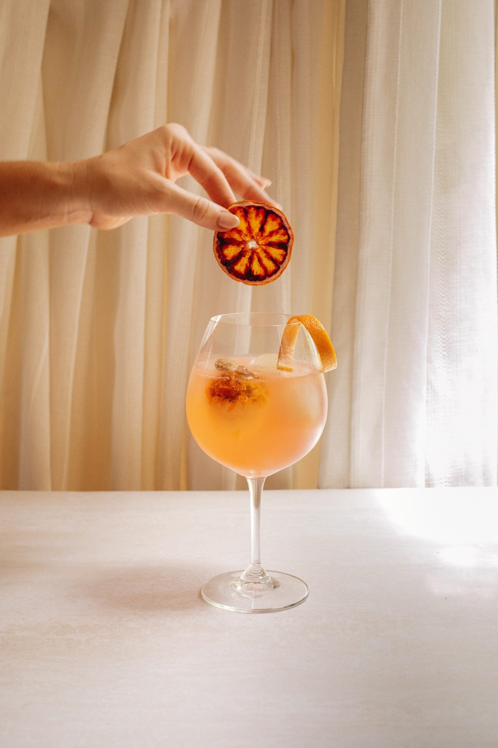 Close-up of orange cocktail with a hand dropping a dried orange into the glass.