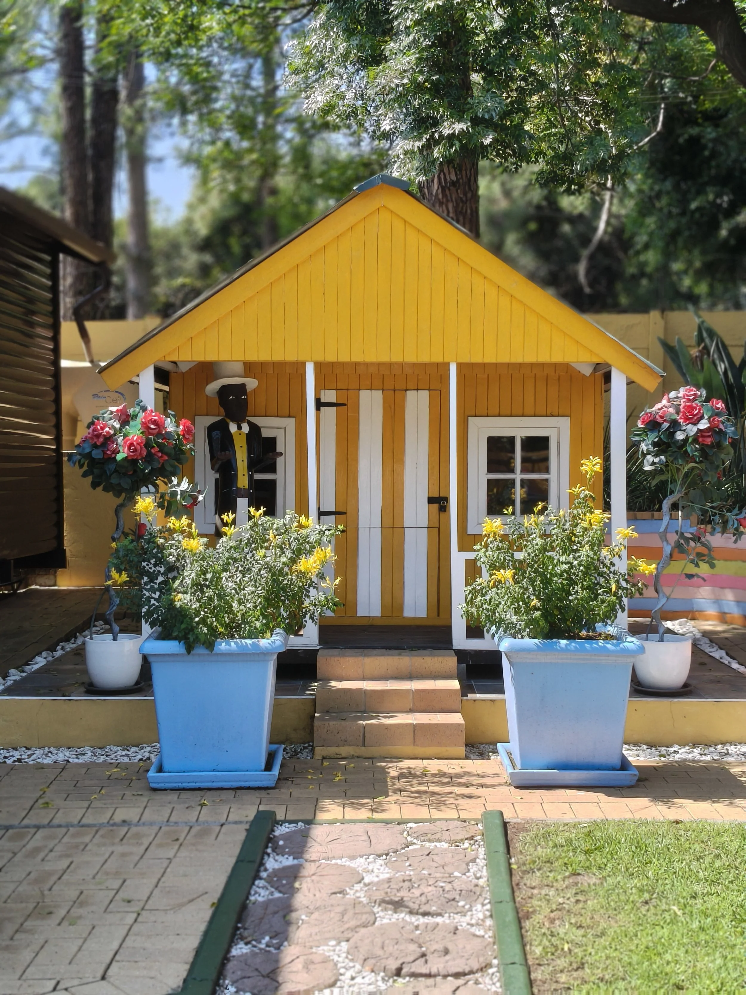 Small yellow house with white trim and a front door, surrounded by potted flowers and plants, with trees in the background.
