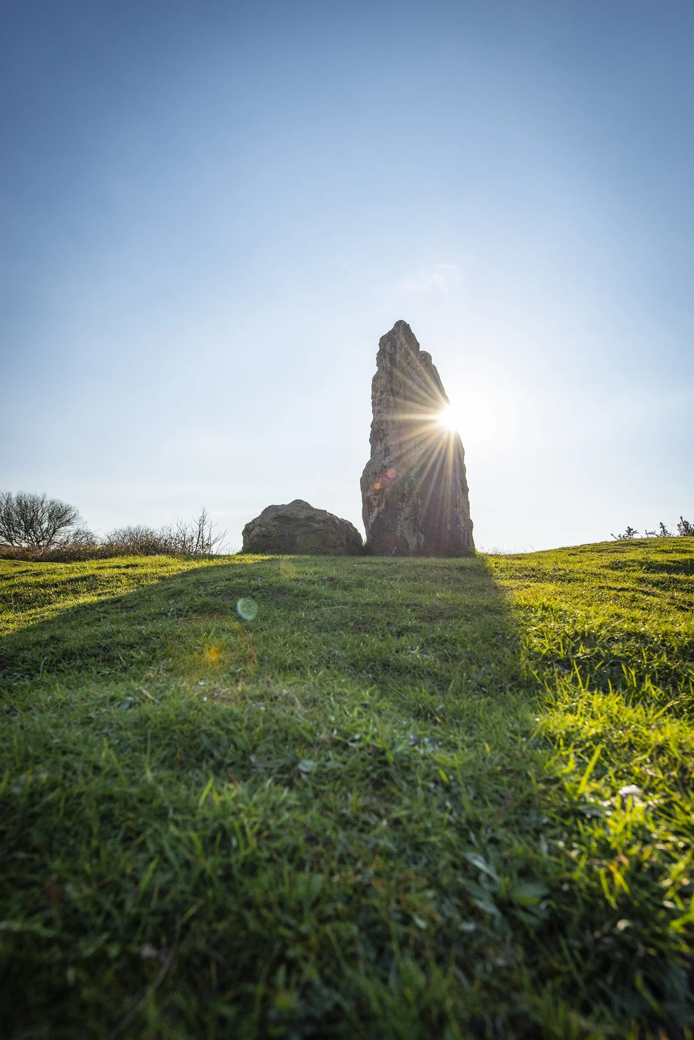 Sun shining behind a tall, narrow stone standing upright on a grassy hill, with sunlight creating a starburst effect.