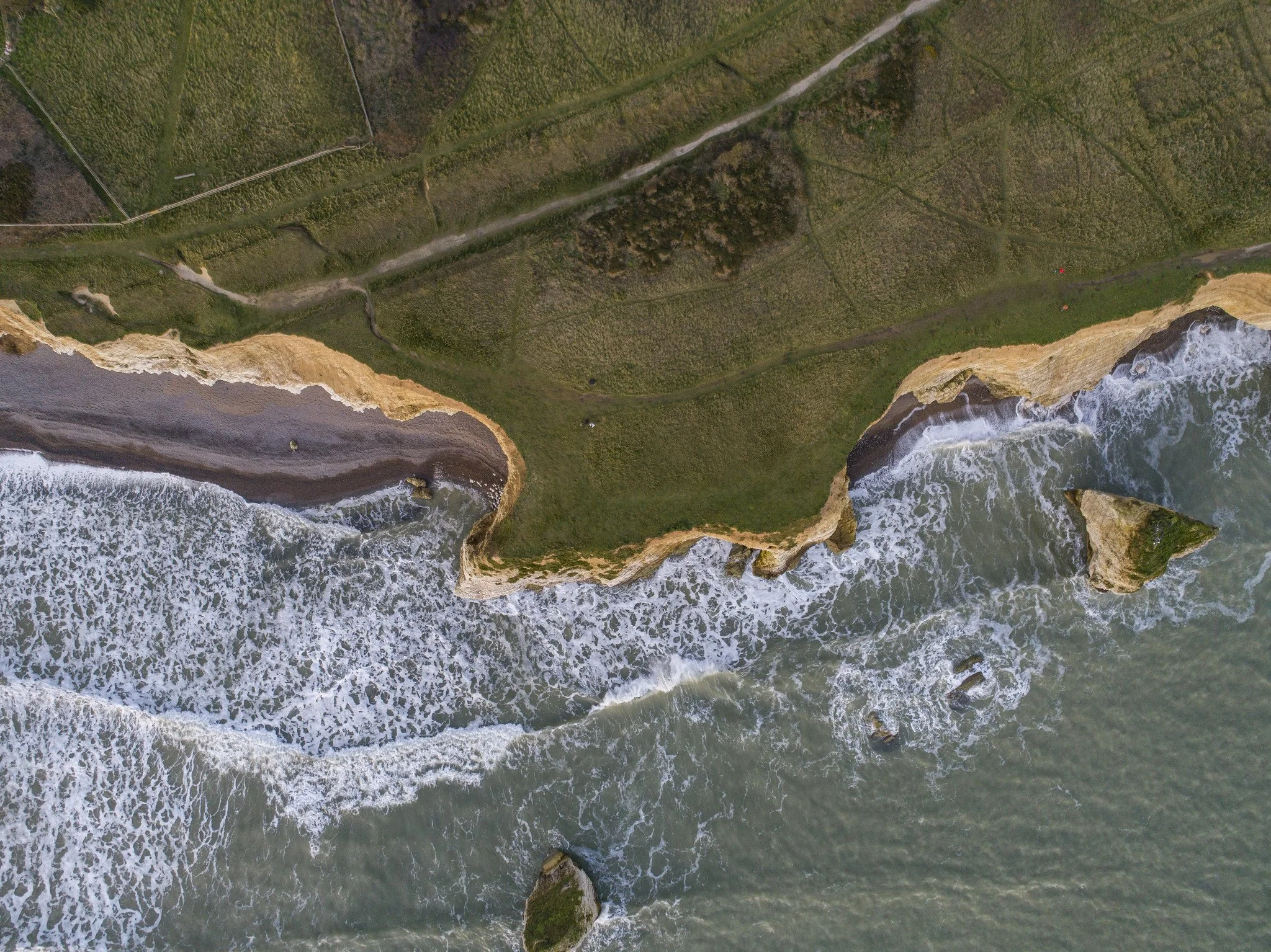Aerial view of rugged cliffs and green grassy land along a shoreline with waves crashing against the rocks.