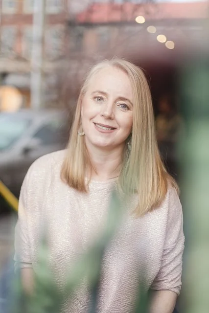 A smiling woman with long blonde hair standing outdoors in an urban setting, with blurred background and some greenery in the foreground.
