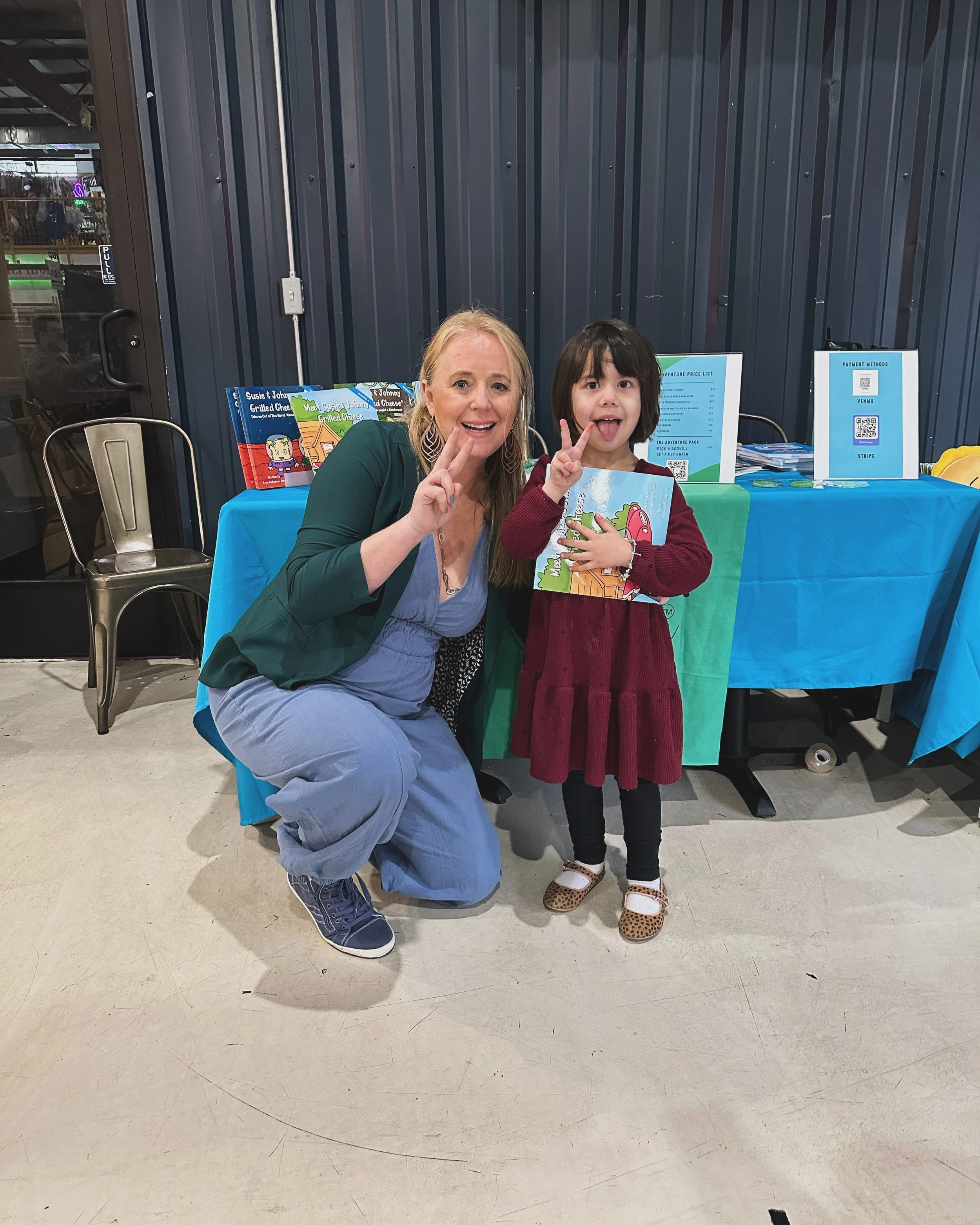 A woman and a young girl posing together at a table with books and promotional materials, both making peace signs and smiling.