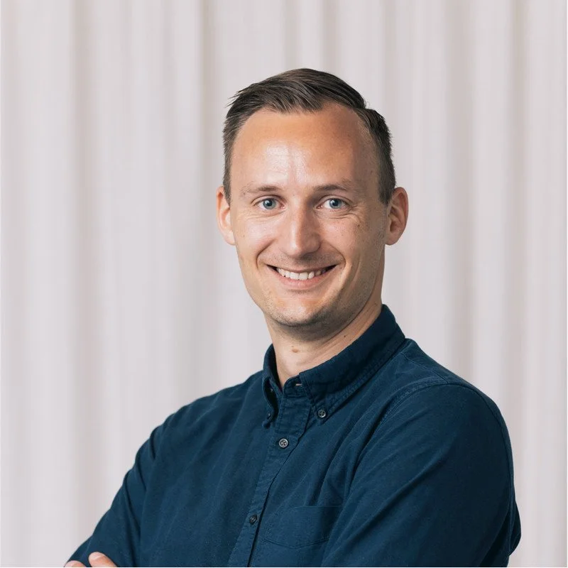 Man smiling, wearing a dark blue shirt, standing in front of a white curtain backdrop.