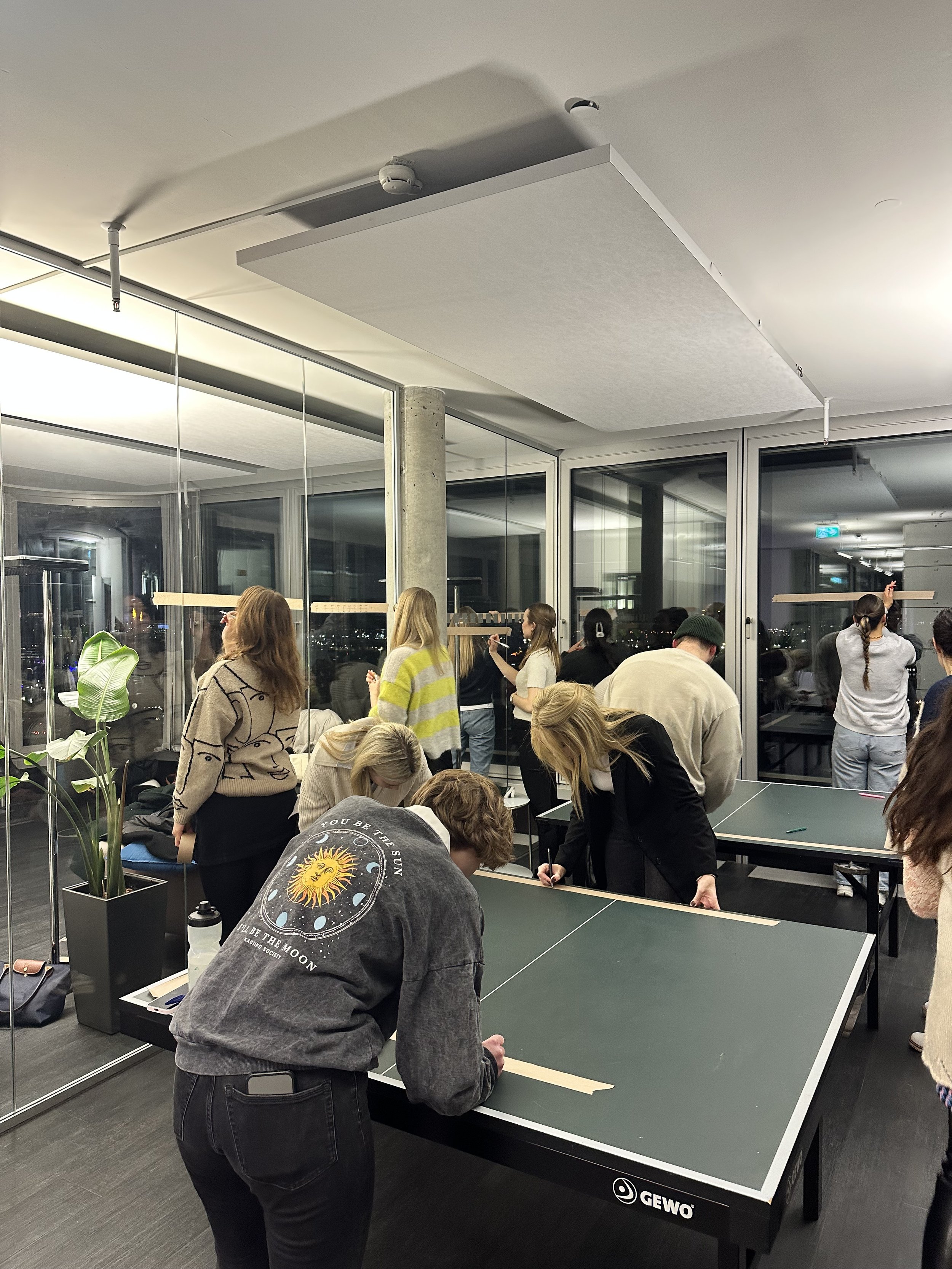 People gathered around a table in an office setting, some standing and others bending over, engaging in a group activity. Large glass windows and overhead lighting are visible. A table tennis table is being used for the activity. Indoor plant and var