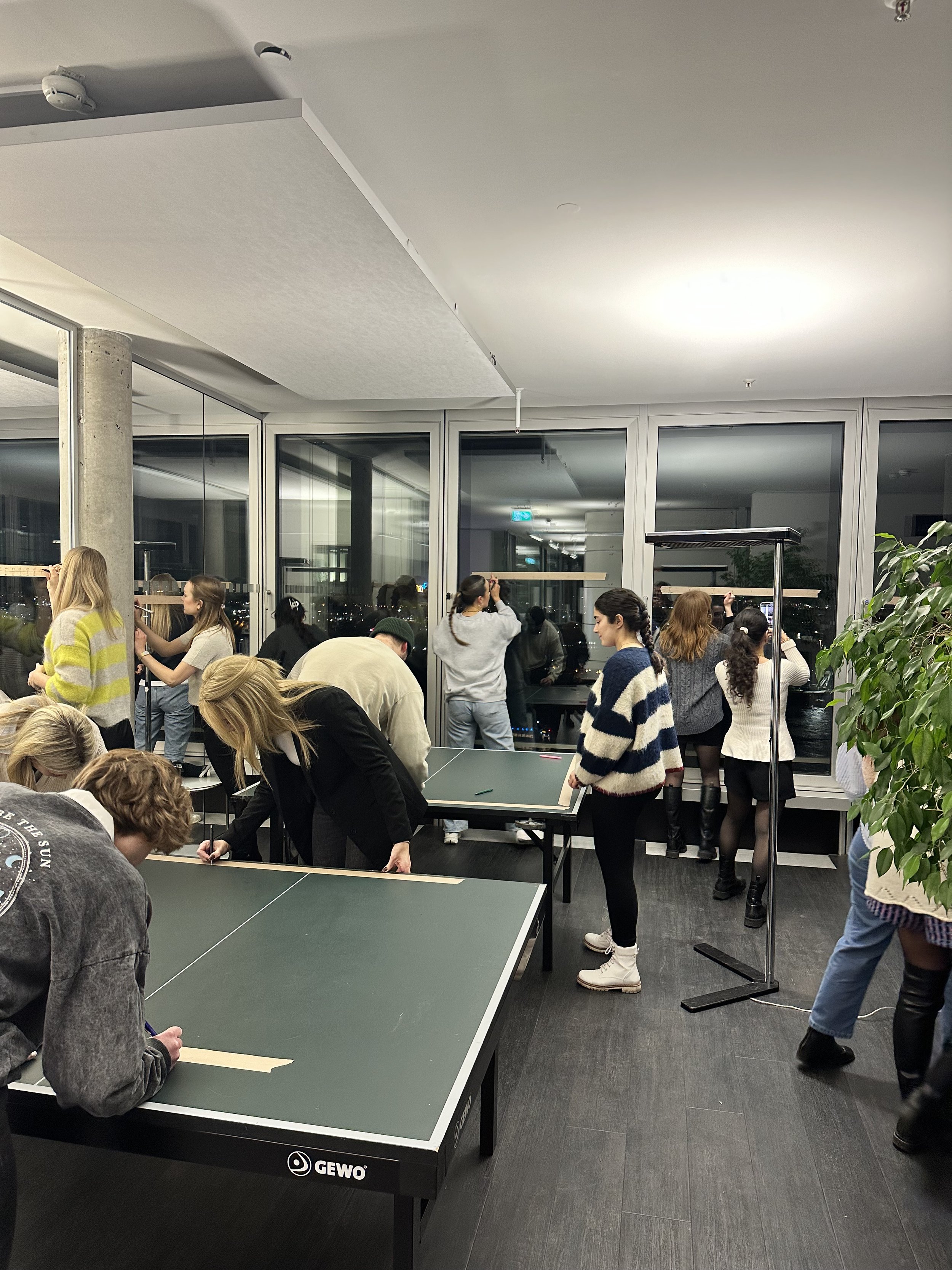People gathered in a room with large windows, using pens on two table tennis tables.