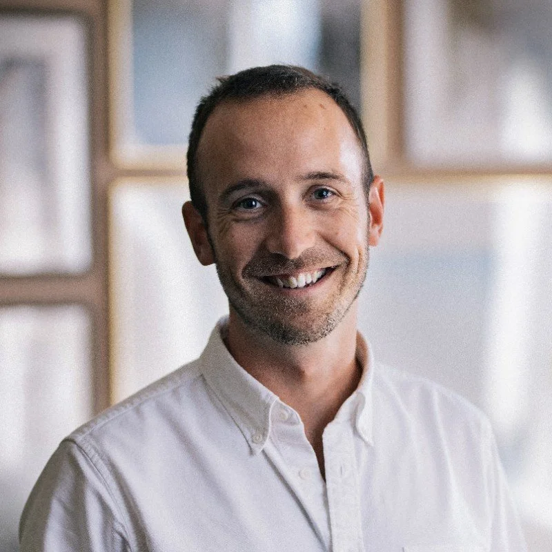 Smiling person wearing a white button-up shirt, standing indoors against a blurred background.