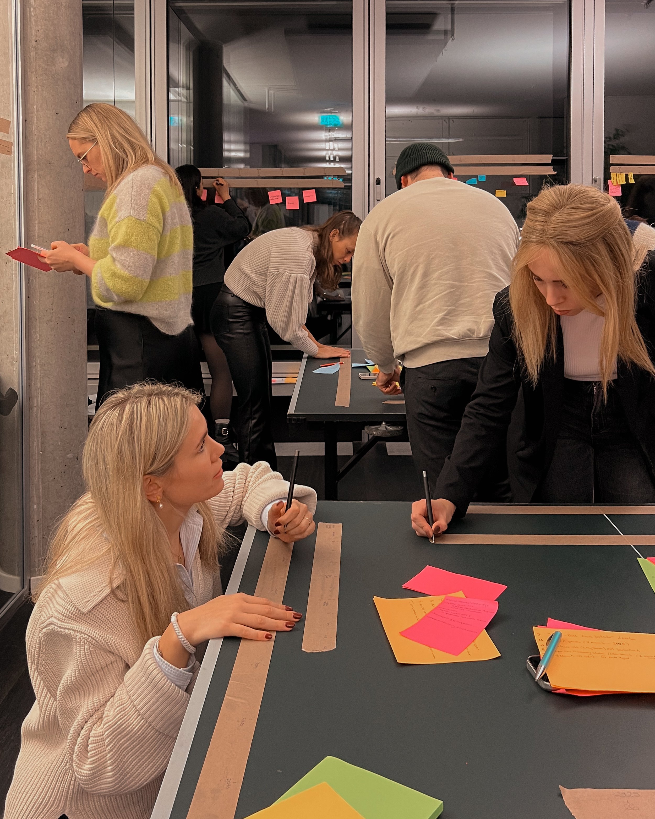 People engaged in a collaborative office activity at a table, writing on colorful papers and using sticky notes. Some participants are standing, while others are sitting, working on tasks together in an indoor setting.