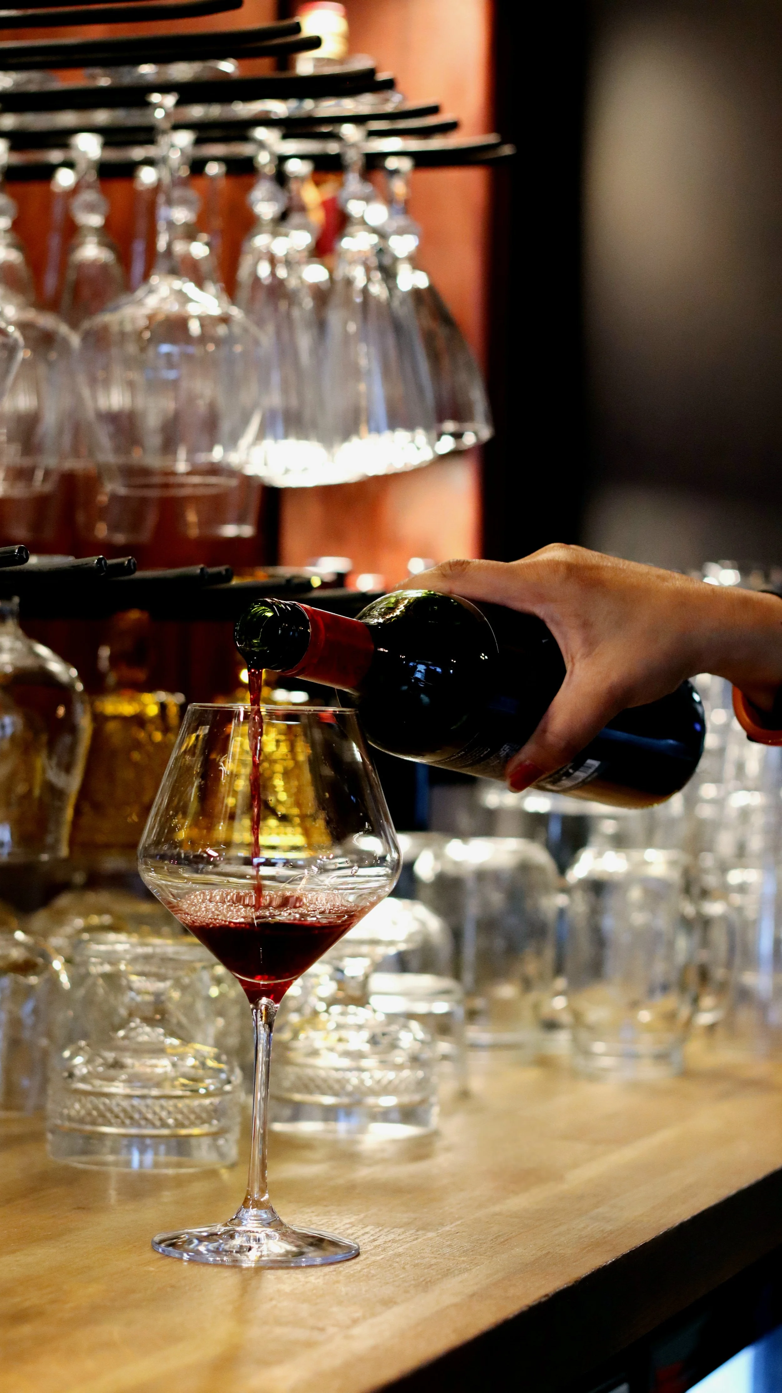 En hand häller rött vin i ett vinglas på en bardisk, med upphängda glas i bakgrunden.
