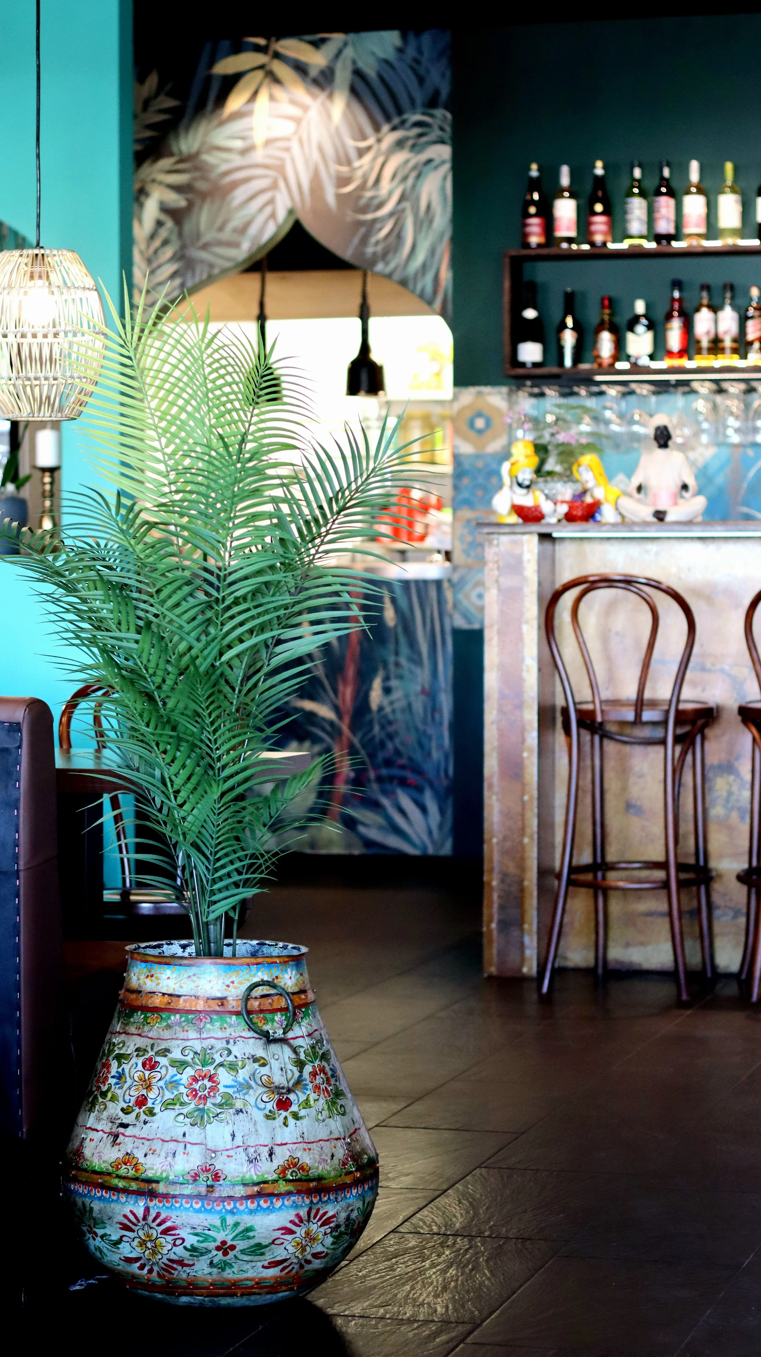 Interiör av en restaurang eller bar med en dekorativ färgglad kruka innehållande en grön växt. Bakgrunden har en bar med stolar och hyllor fyllda med flaskor. Väggarna är dekorerade med växtmotiv.