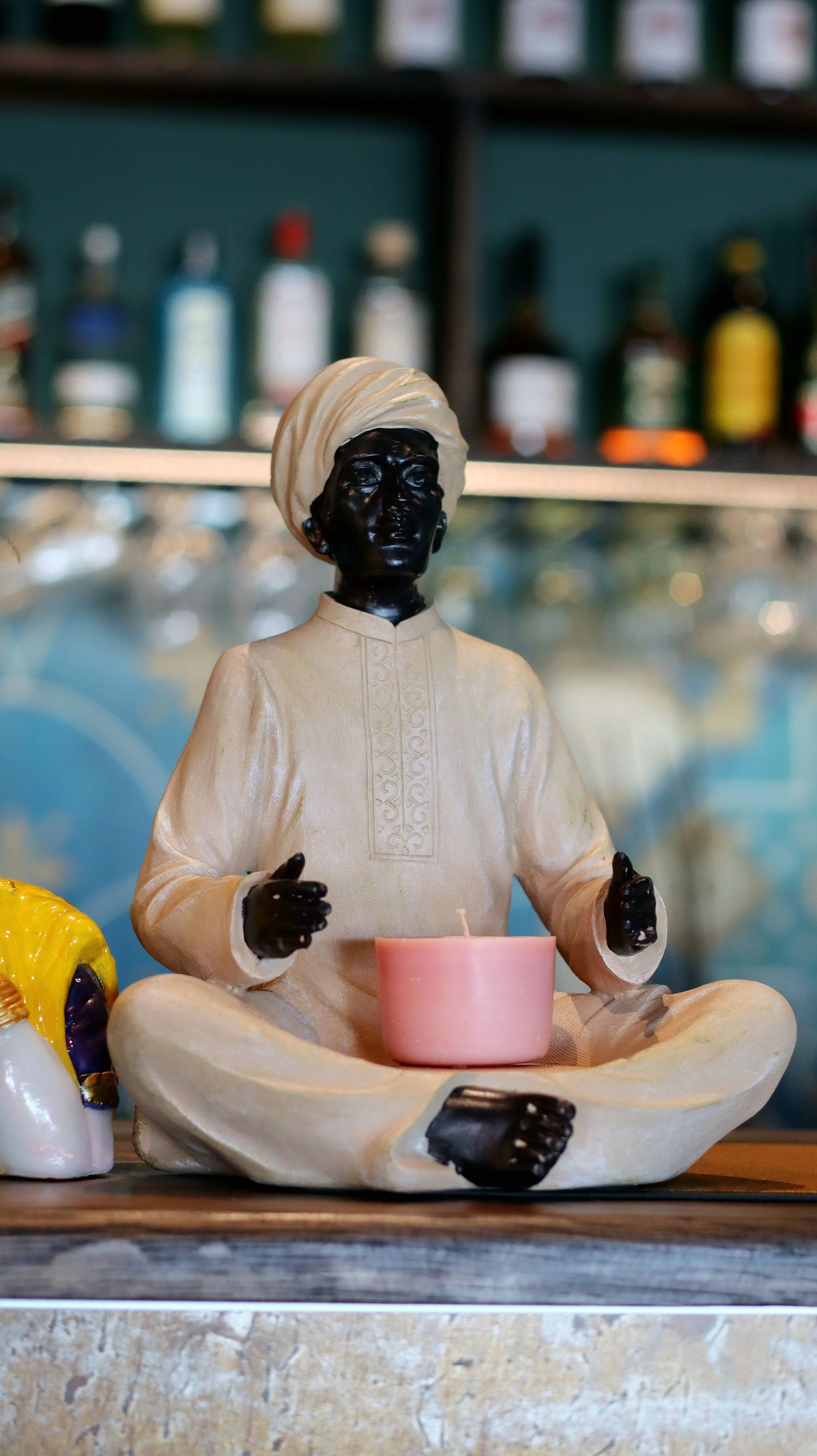 Staty av sittande person i traditionella kläder med ljus, inomhus, med flaskor i bakgrunden.