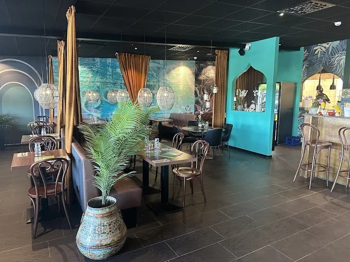 En inredning av en restaurang med sittplatser, trästolar och bord. Väggarna är turkosa med dekorativa mönster, och det finns en stor krukväxt. Ljuskronor hänger från taket. Det finns också en bar till höger.