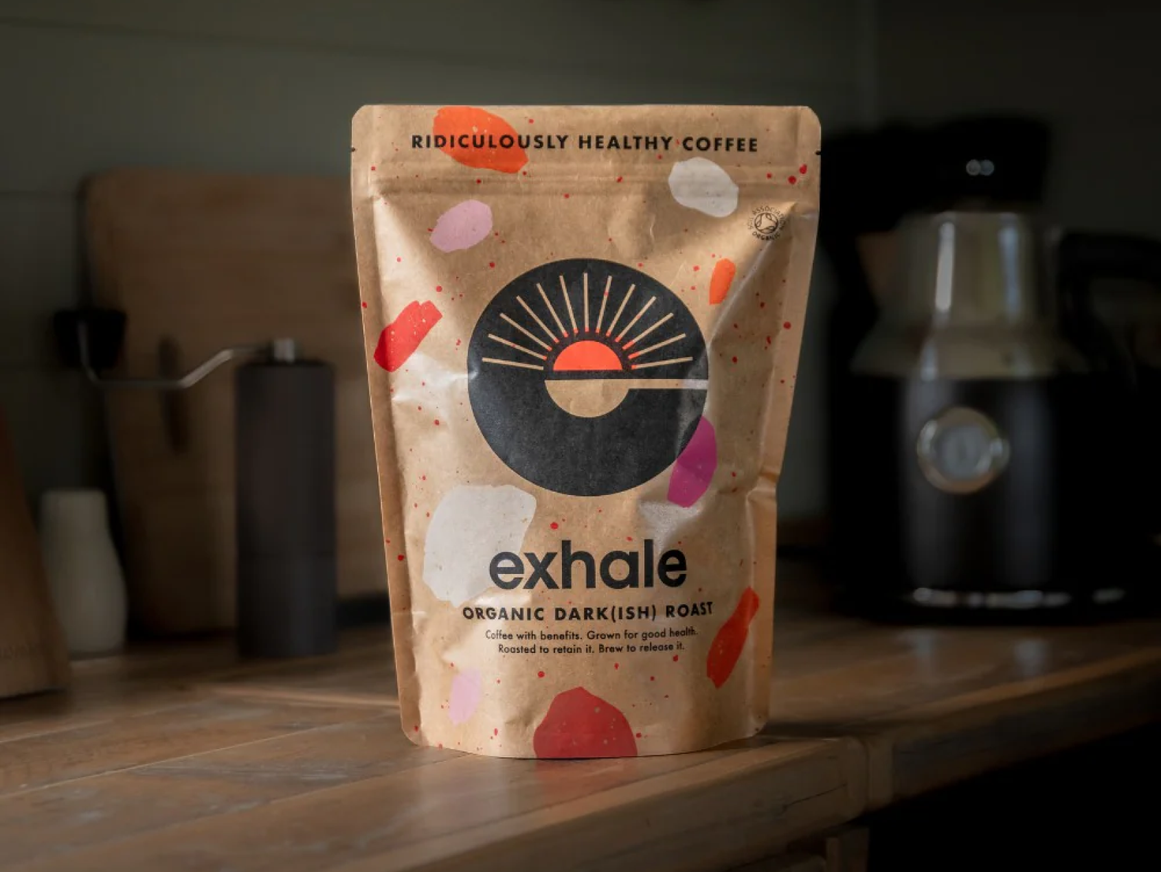 A beige paper coffee bag labeled 'exhale' with colorful abstract shapes, sitting on a wooden countertop with a coffee grinder in the background.