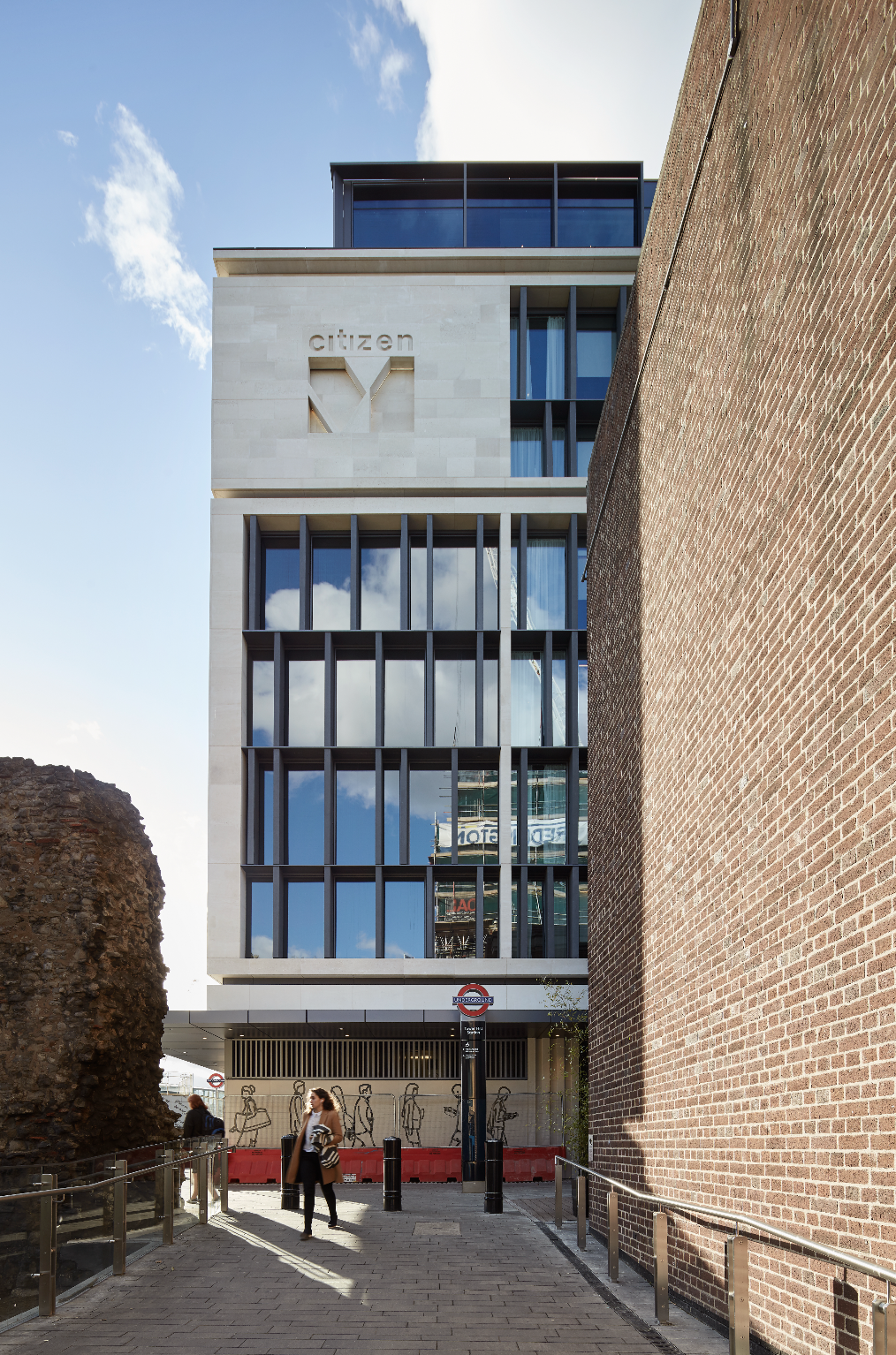 Modern building with glass windows and a "Citizen" sign, located between brick and historic ruins, with pedestrians walking nearby under a partly cloudy sky.