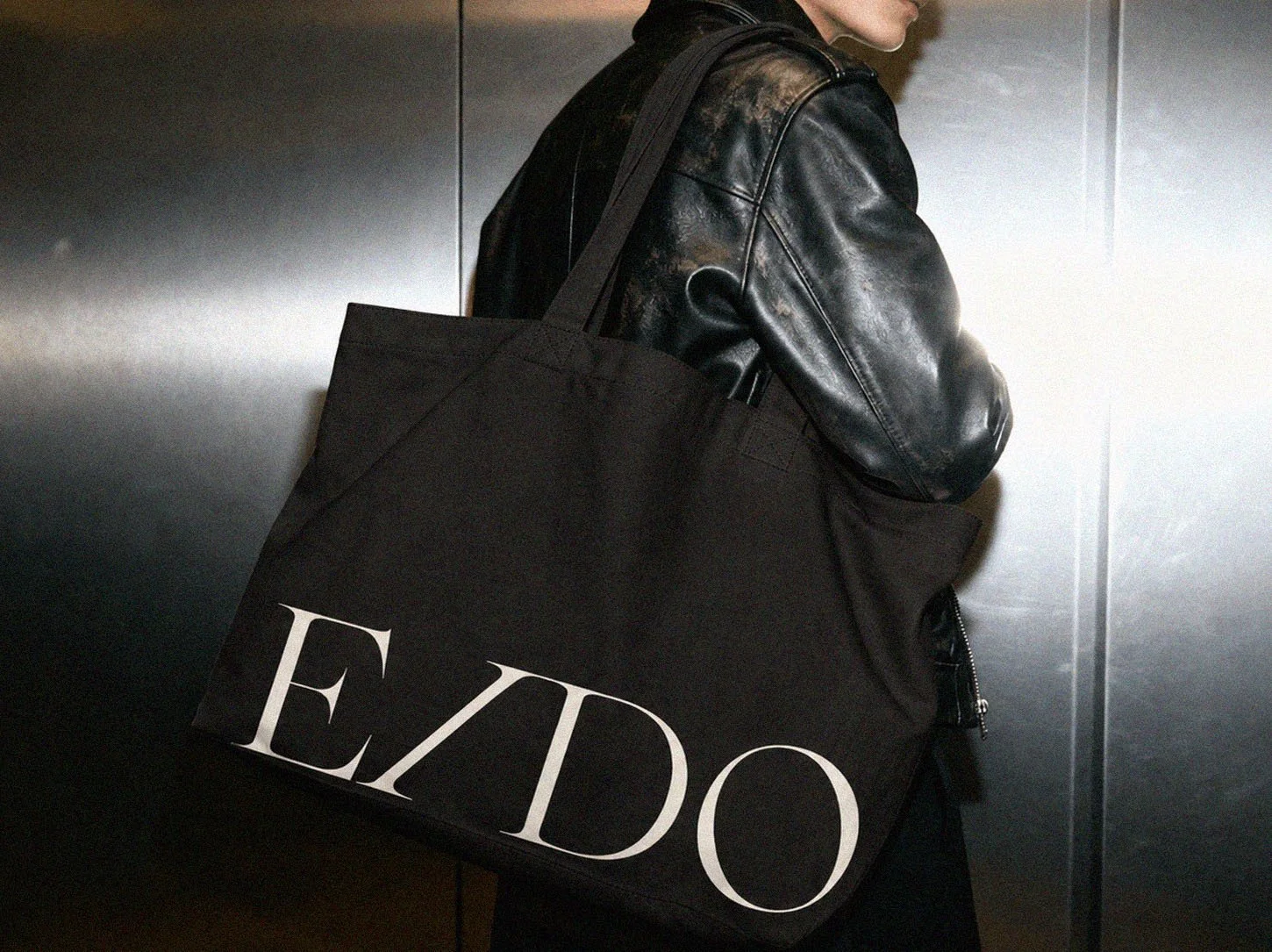 Person wearing a black leather jacket carrying a large black tote bag with the word 'ELDO' in large white letters, in an elevator with metallic walls.