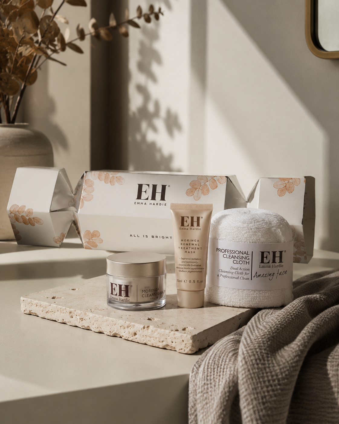 Skincare products displayed on a white surface, including a box, a jar, a tube, and a cleansing cloth, with decorative plant and shadow in the background.