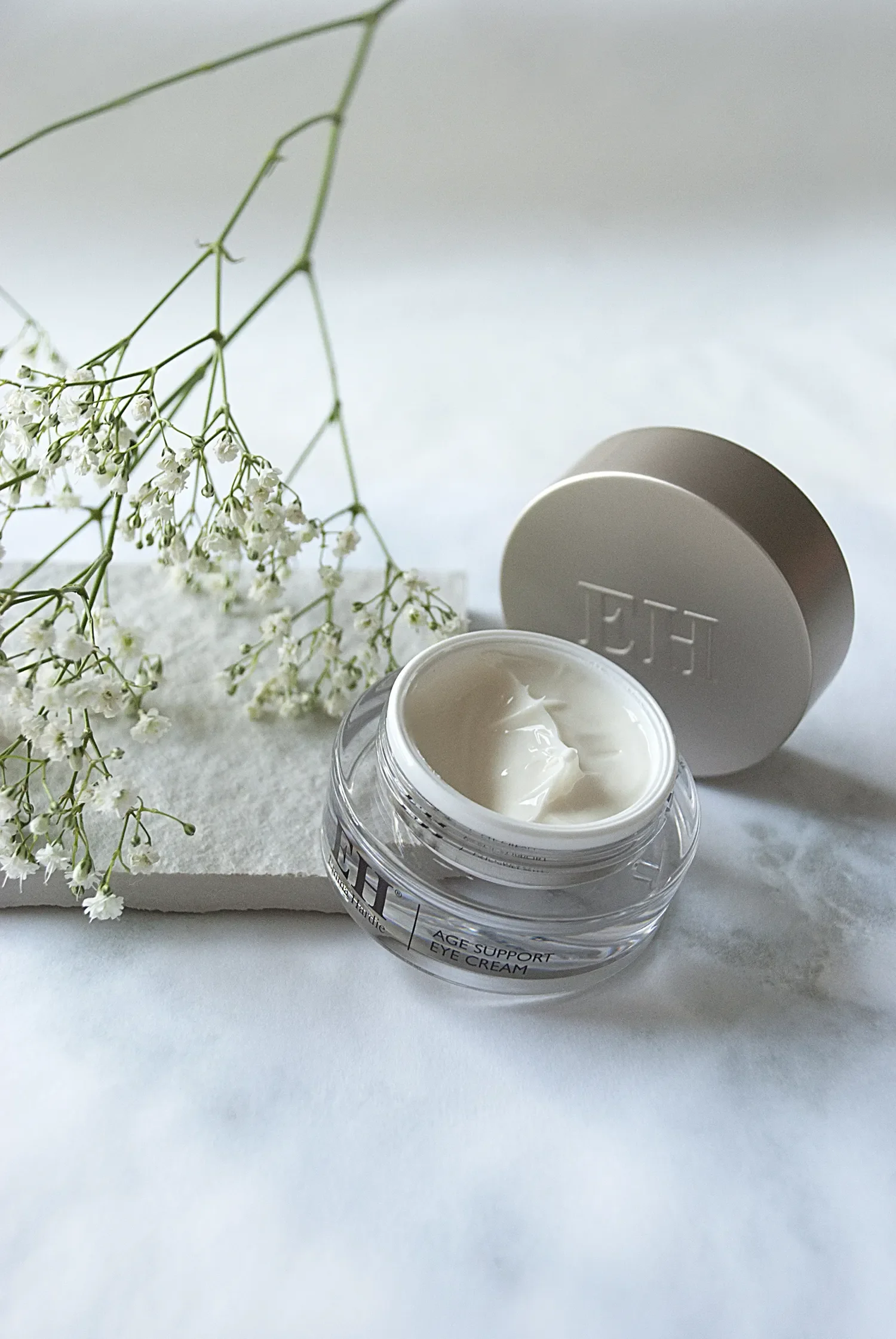 An open container of age support eye cream with a white lid beside a sprig of small white flowers on a white surface.