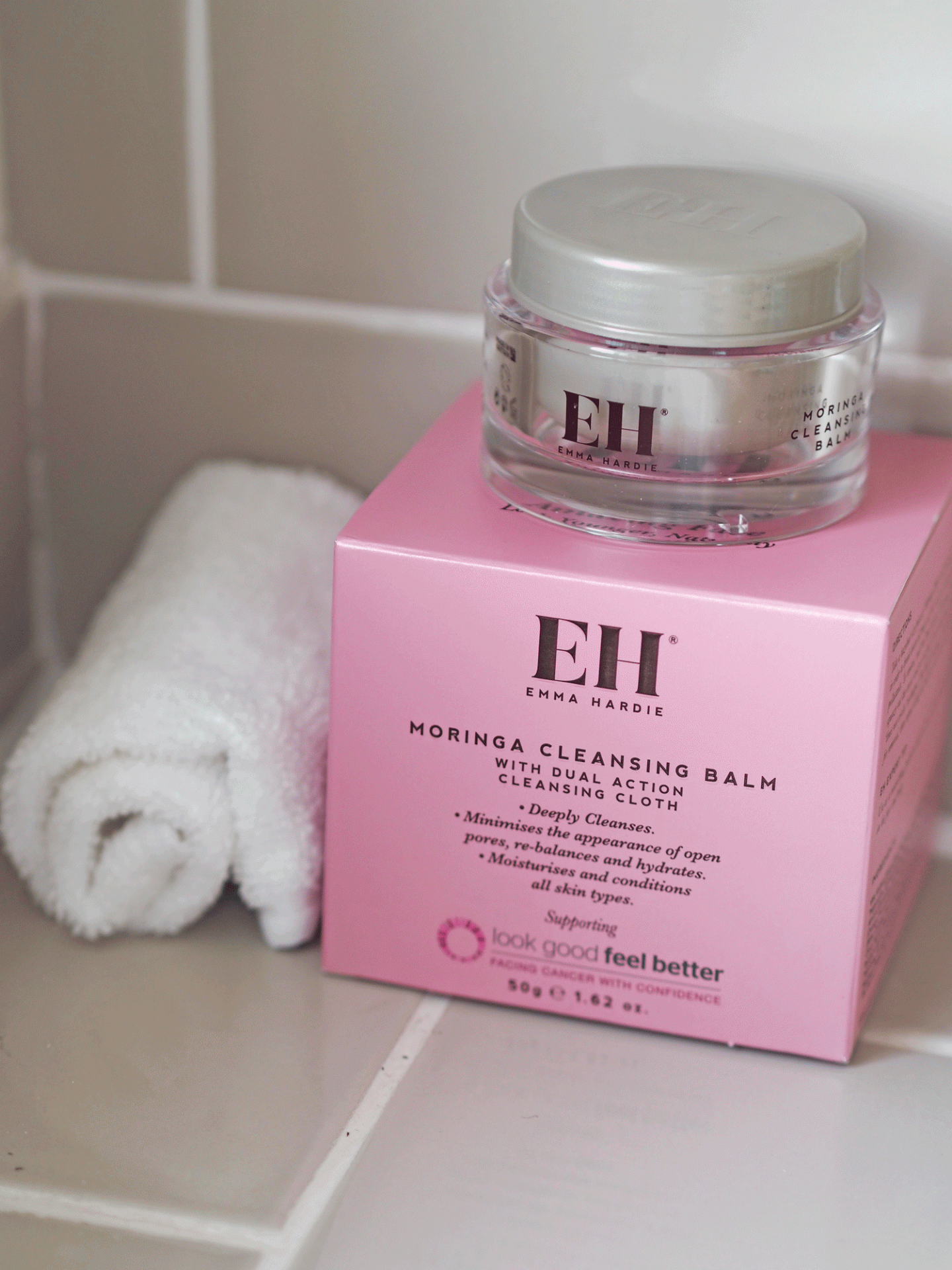 A pink box of Emma Hardie MORINGA CLEANSING BALM with a glass jar of the same product on top, placed beside a rolled white towel on a bathroom countertop.