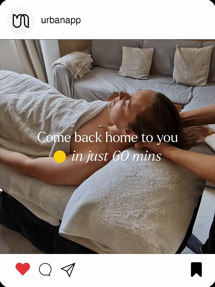 A woman lying on a massage table with her head resting on a pillow, receiving a massage in a cozy living room setting with a sofa and pillows in the background.