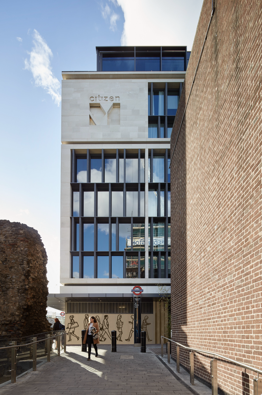 Modern building with glass windows and the word 'citizen' on top, located between a brick wall and ancient ruins, with people walking on the sidewalk in front, under a partly cloudy sky.