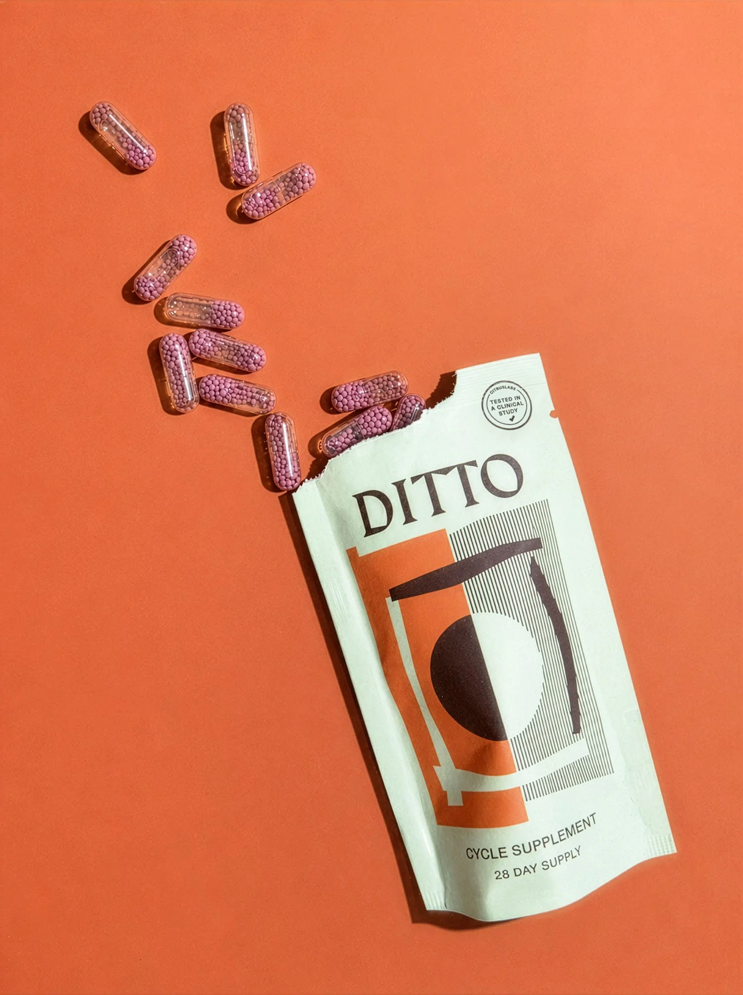 A partially opened white package with black text and graphic design, labeled 'DITTO' and 'Cycle Supplement' on a red background, with several pink and purple capsules spilled out.
