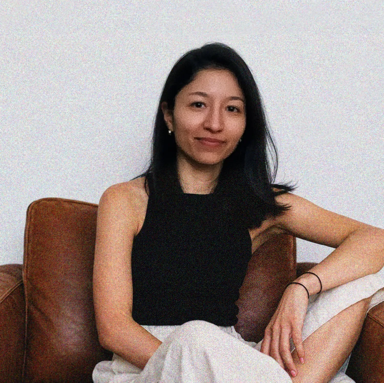 A woman with shoulder-length black hair sitting on a brown couch, wearing a black sleeveless top and white pants, against a plain white wall.