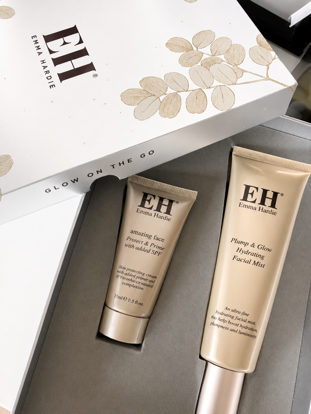 Emma Hardie skincare products including a tube of Plump & Glow Hydrating Facial Mist and a smaller tube of Amazing Face Protect & Prime on a gray surface, with a white box with gold leaf design and the text 'Glow on the Go' partially visible.