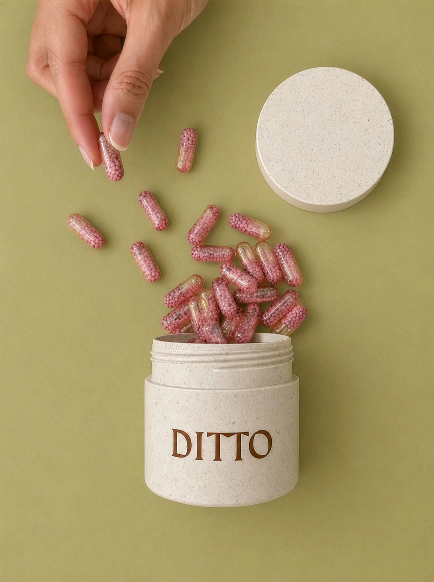 A hand dropping pink capsule-shaped pills into a container labeled 'DITO' with a matching lid nearby, on a green background.