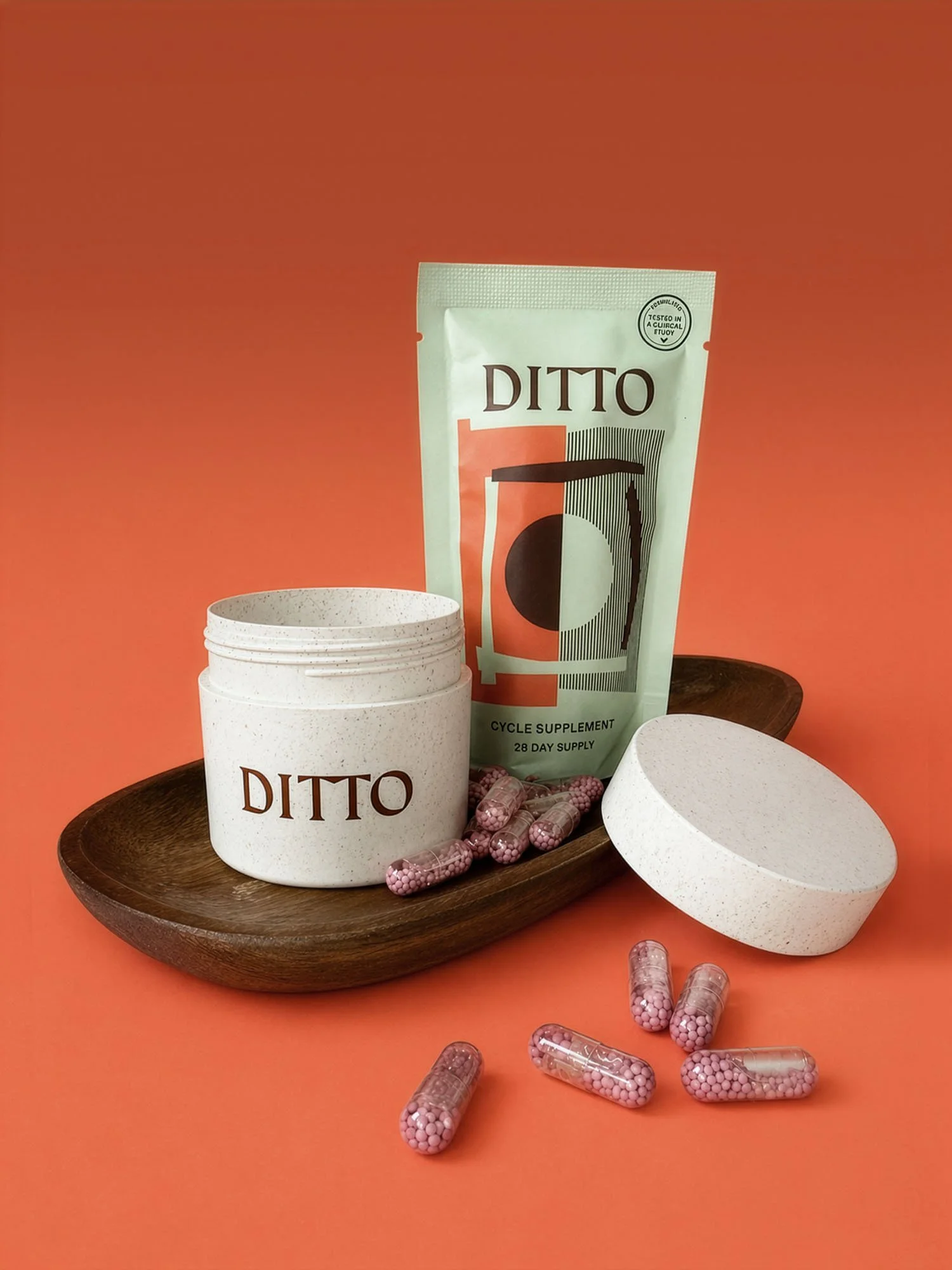 A package of Ditto cycle supplement standing on a wooden tray surrounded by open white containers and scattered pink and white capsules on an orange background.