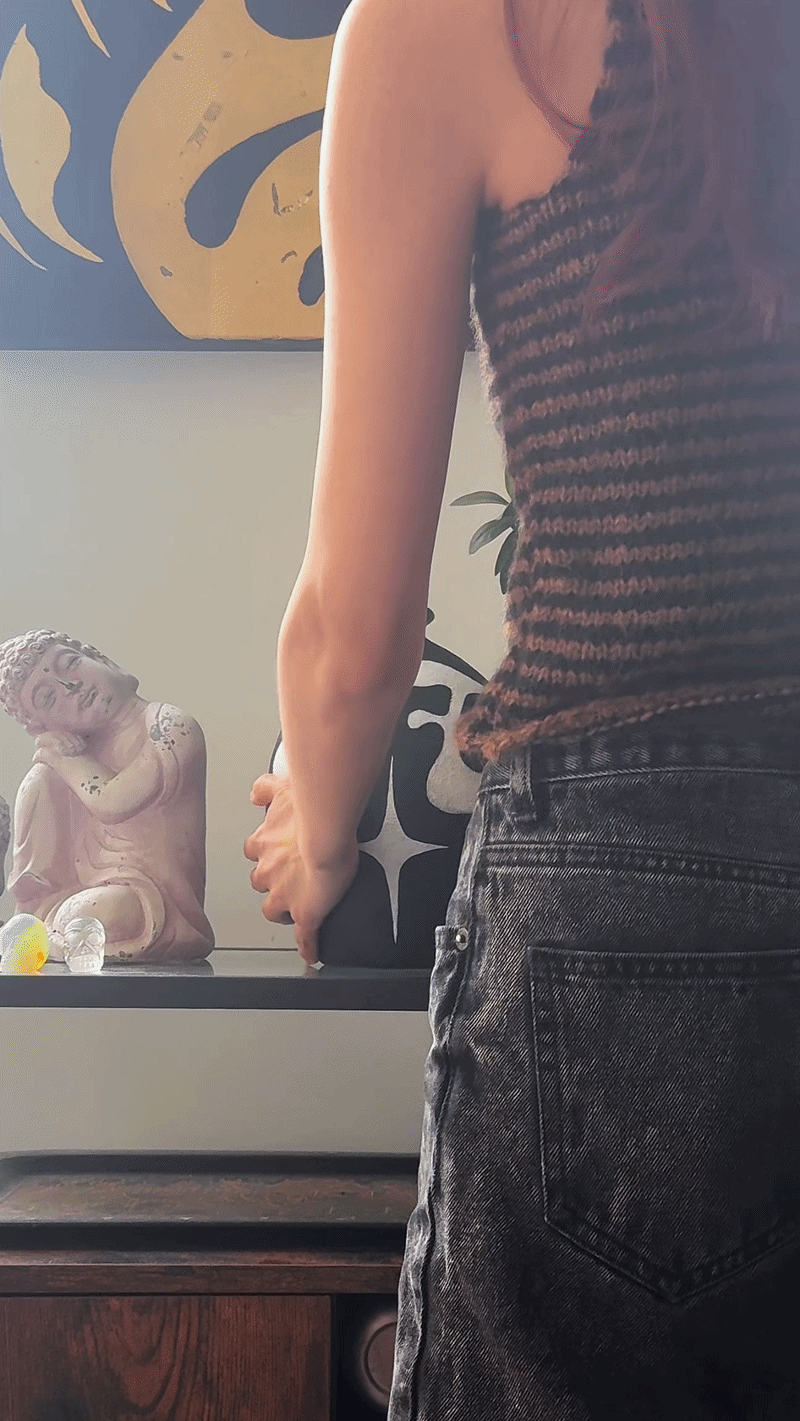 A person in a sleeveless striped top and black jeans standing next to a table with a Buddha statue and decorative objects.